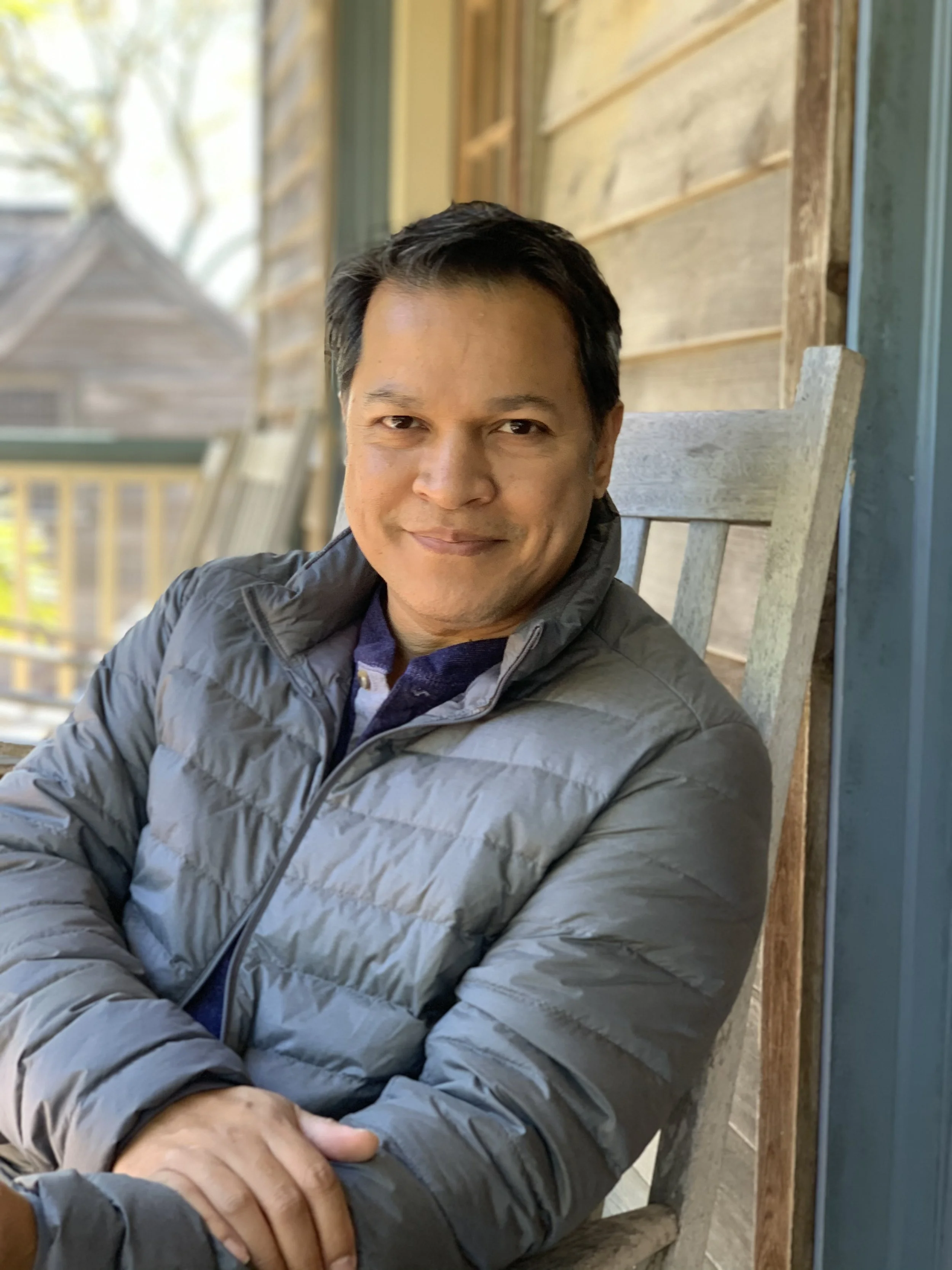 A man sitting outdoors on a wooden chair, wearing a gray jacket and smiling at the camera with a background of a wooden wall and trees.