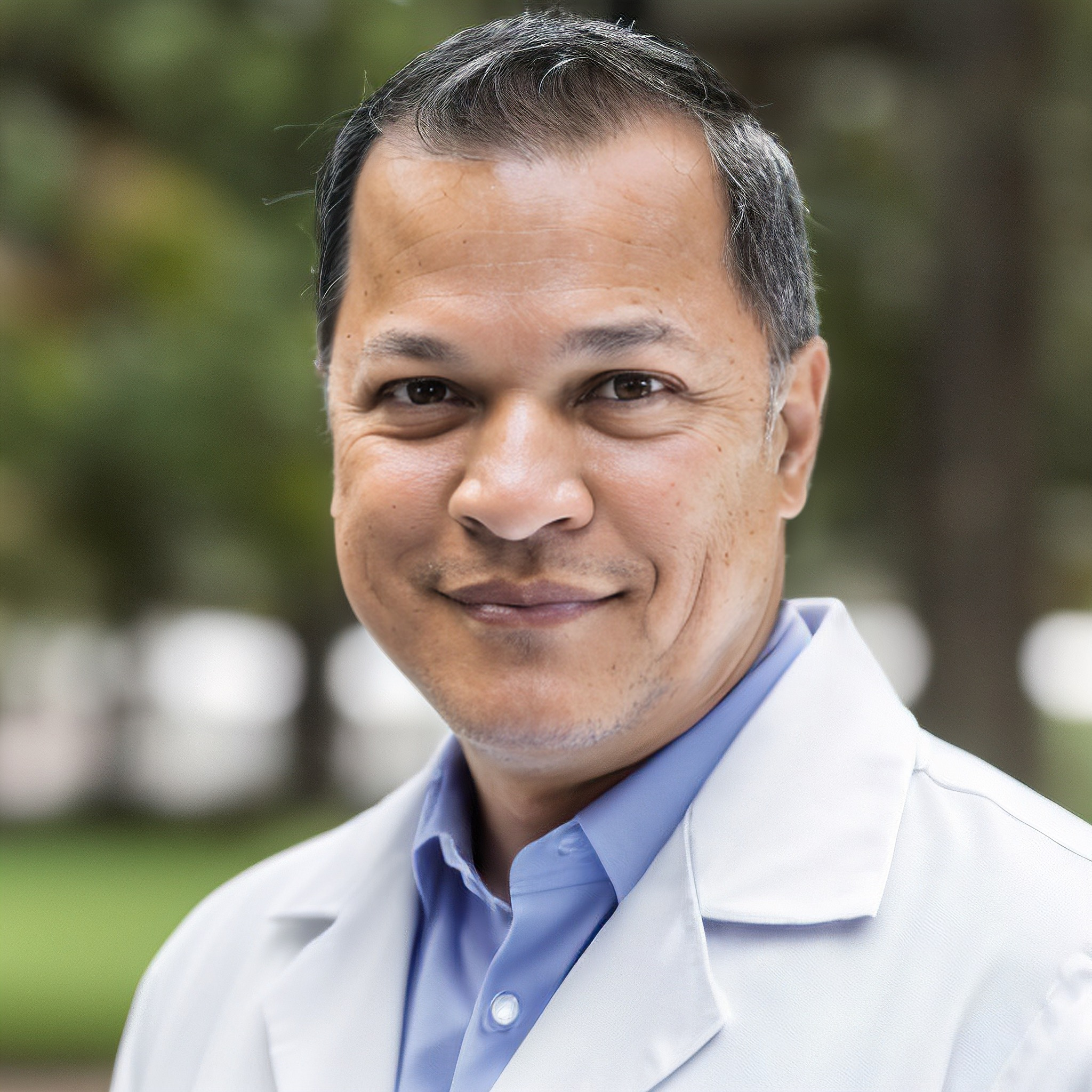 A smiling man in a white lab coat outdoors with trees in the background.