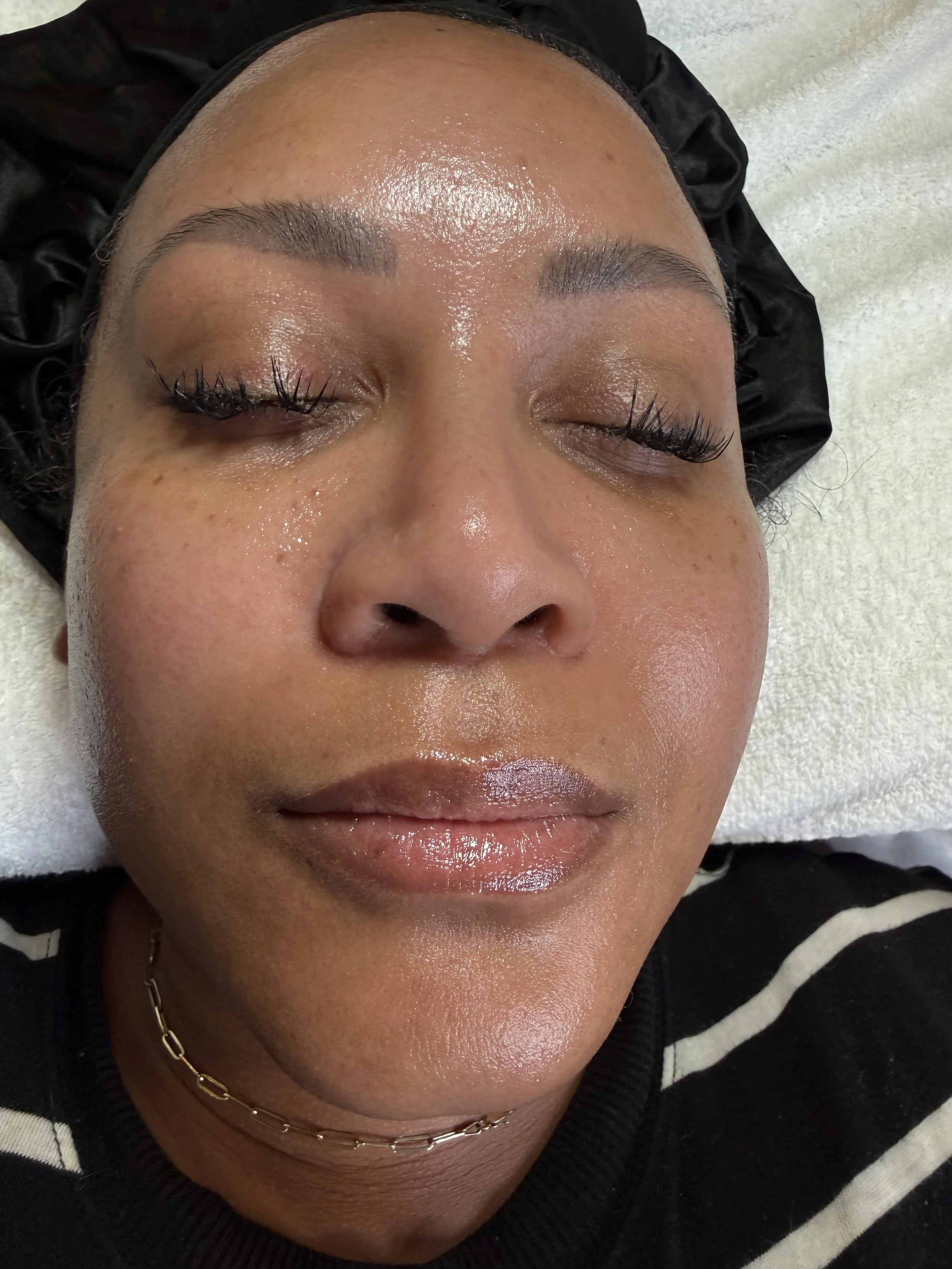 Close-up of a woman's face with closed eyes, wearing long eyelashes, a chain necklace, and a black hoodie with a white stripe, lying on a white towel.