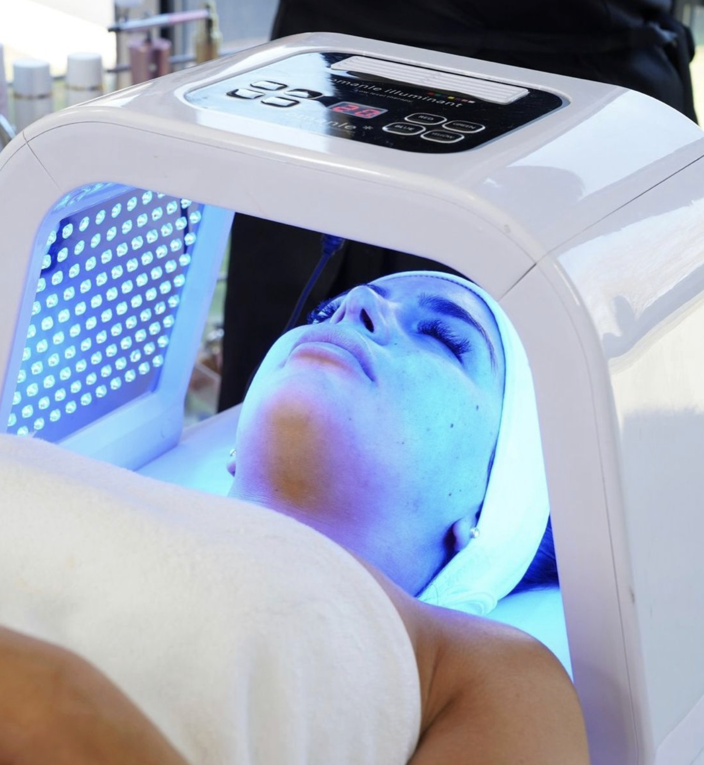 A woman lying under a blue light therapy device with her eyes closed, wearing a white headband. The device has a control panel on top, and the woman appears to be receiving a skincare treatment.