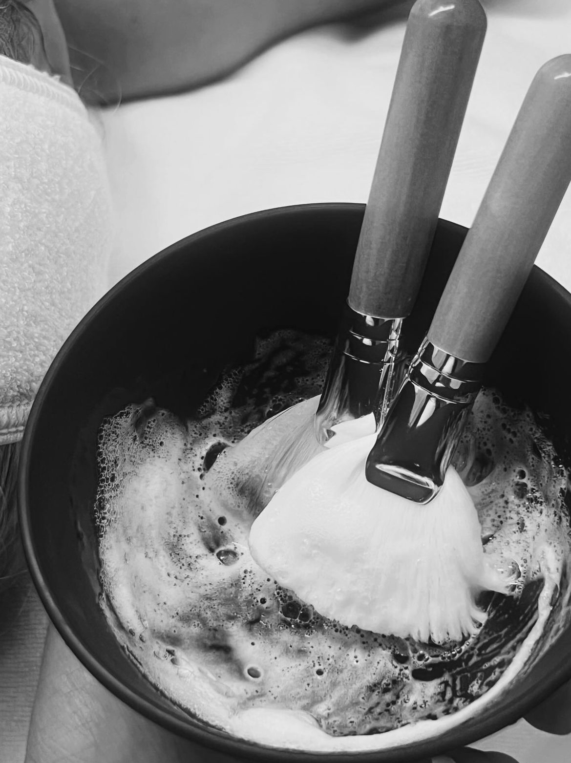 Two shaving brushes in a black bowl filled with foamy shaving cream.