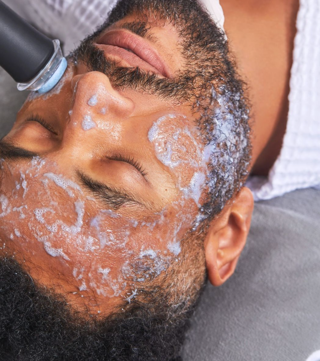 A man with a beard receives a facial cleansing treatment, with a foamy cleanser applied to his face while a beauty technician uses a tool on his forehead.