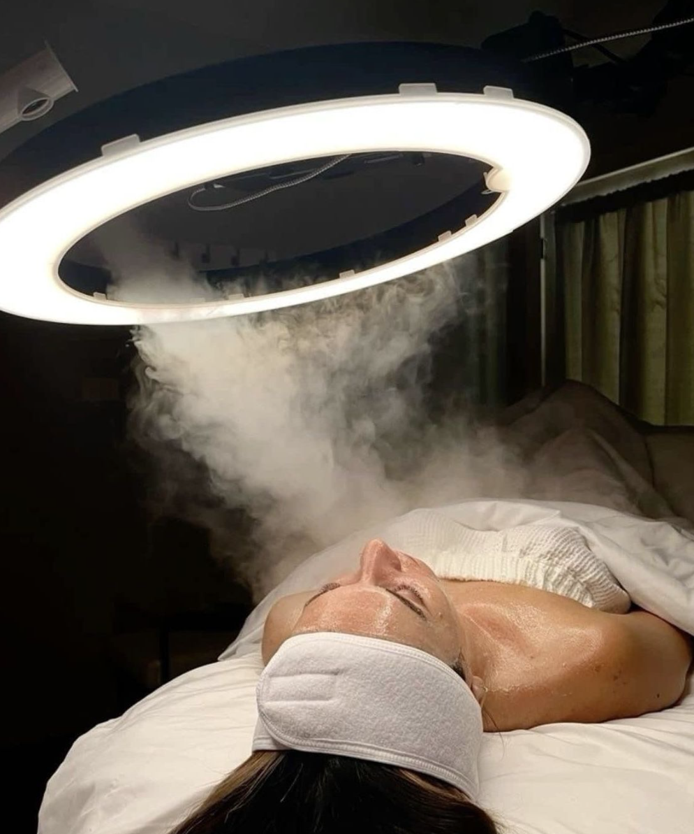 A woman lying in a bed receiving a facial treatment under a circular fluorescent light with steam or vapor emitting from the device.