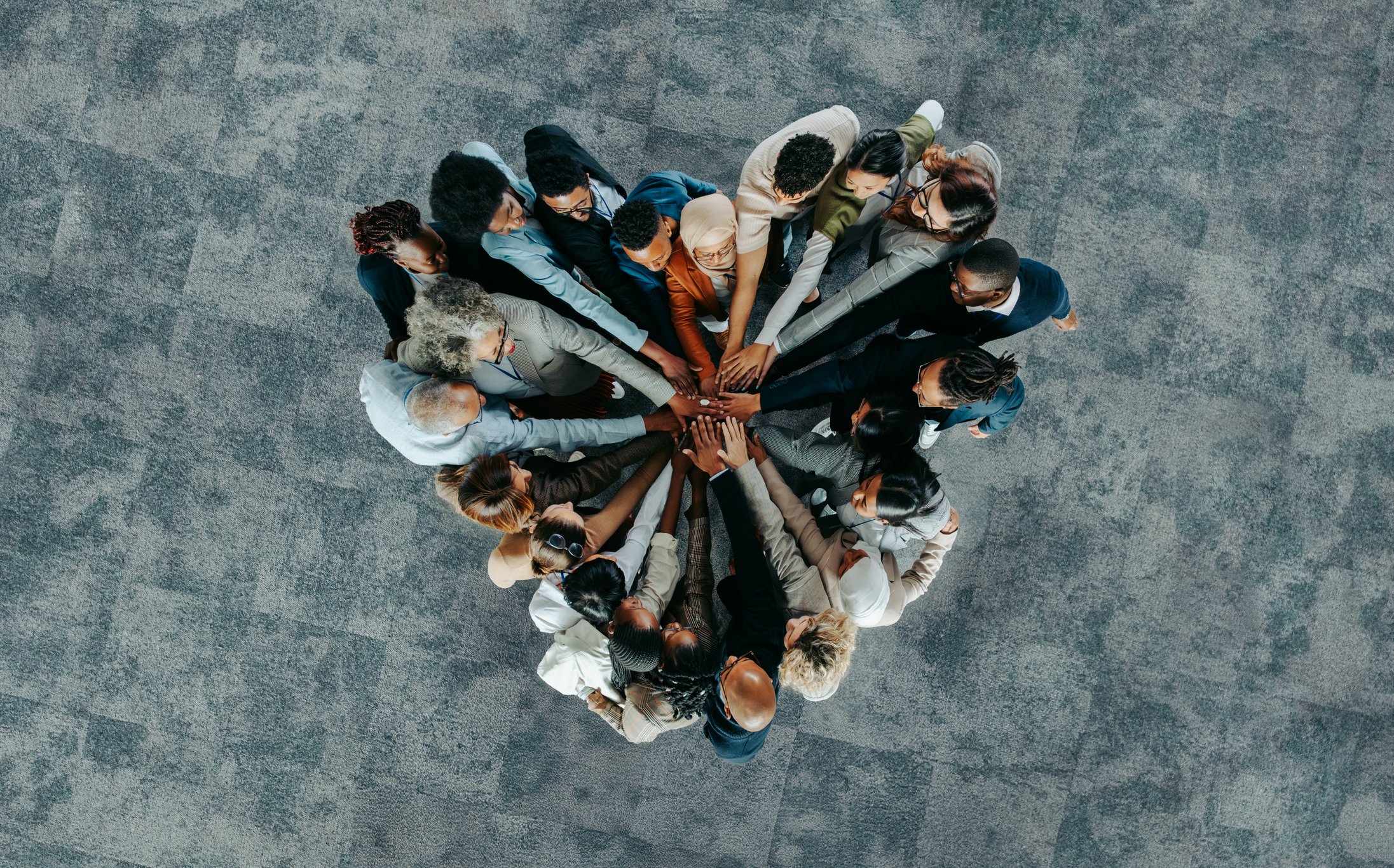 A group of diverse people in business attire standing in a circle with their hands stacked in the center, viewed from above.
