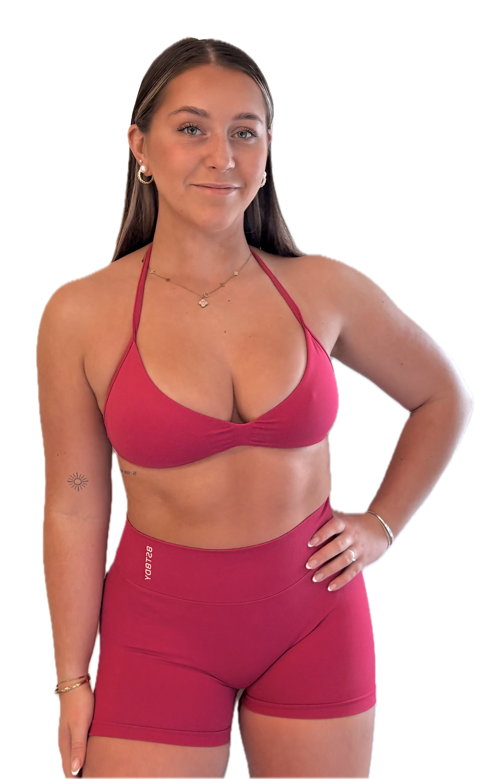 Young woman with long brown hair wearing a pink sports bra and matching pink shorts, standing with her hand on her hip, smiling slightly.