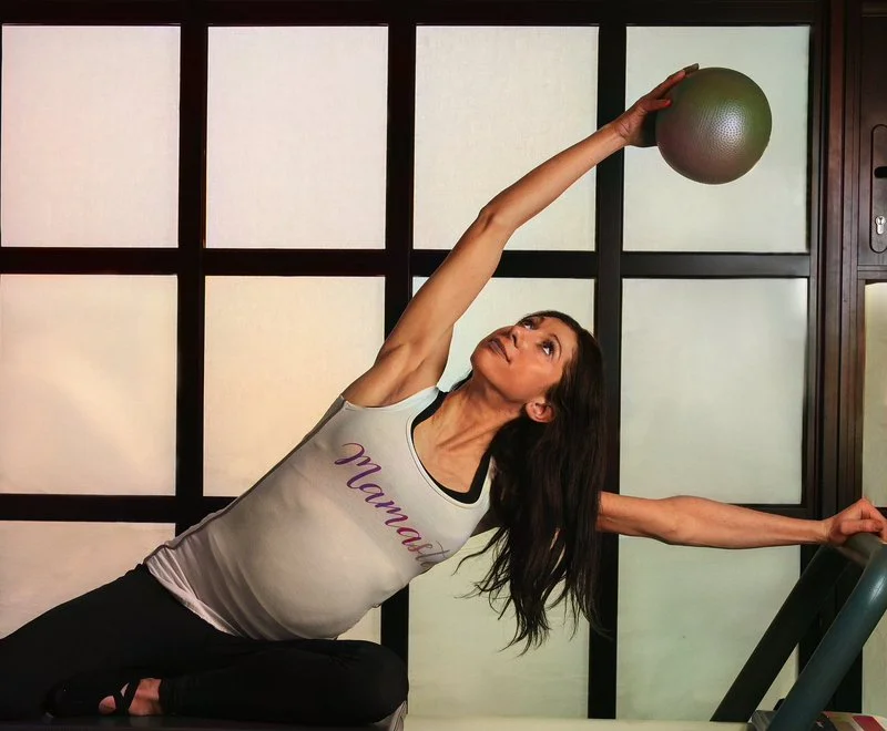 A woman in a gray tank top with the word 'Mama' on it, performing a side stretch exercise while holding a gym ball overhead, seated on a mat in a room with a large grid-patterned window.