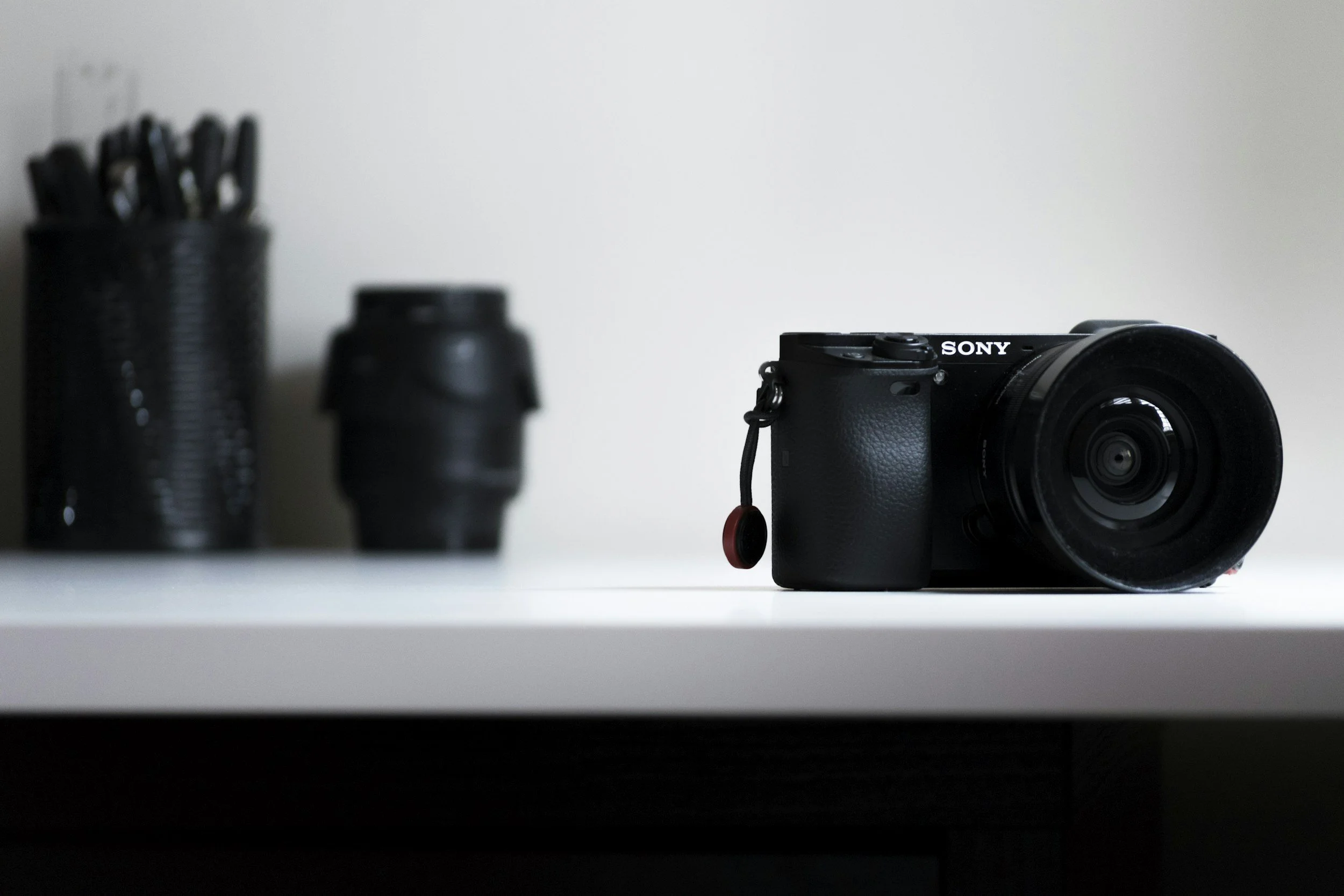 A black digital camera with a large lens sits on a white surface in the foreground, with a blurred container of pens and a black container in the background against a white wall.