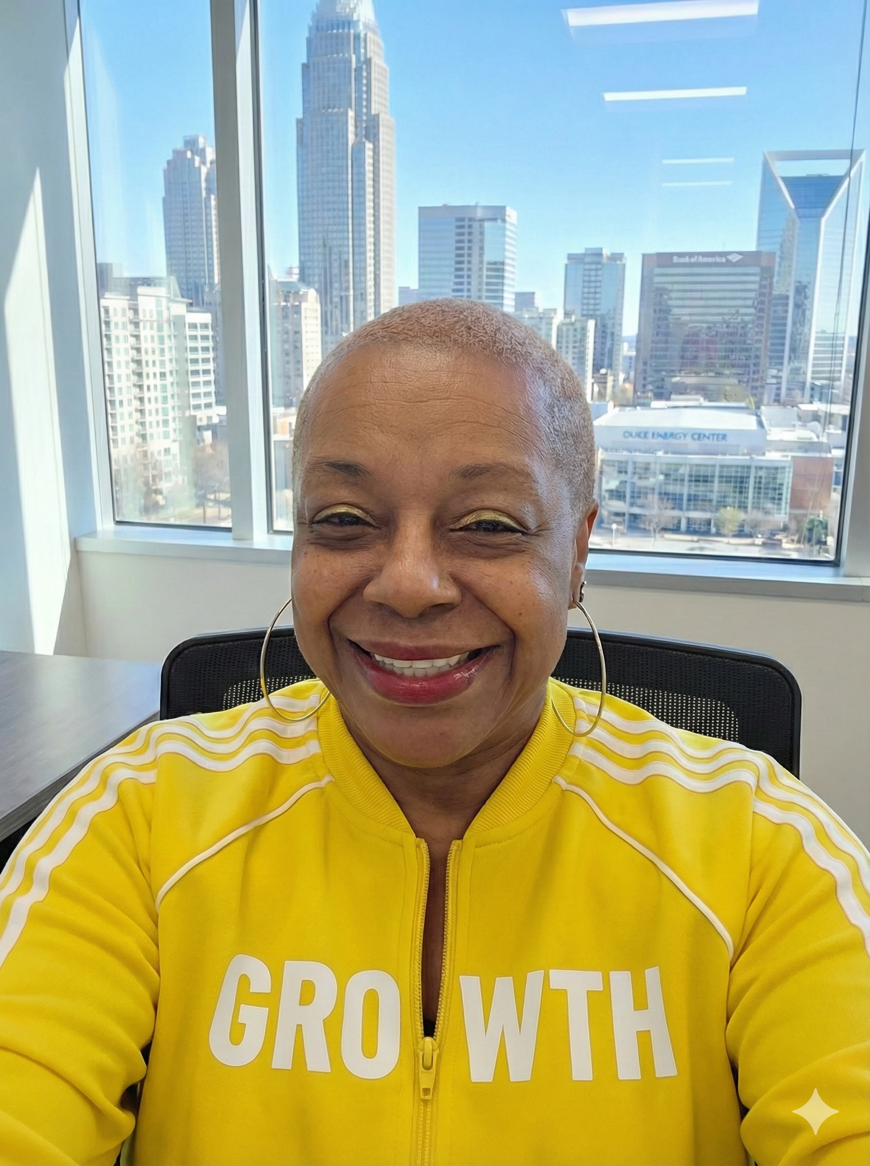 A smiling woman with short hair wearing a yellow jacket with white text that reads 'GROWTH' in an office with large window and cityscape view.