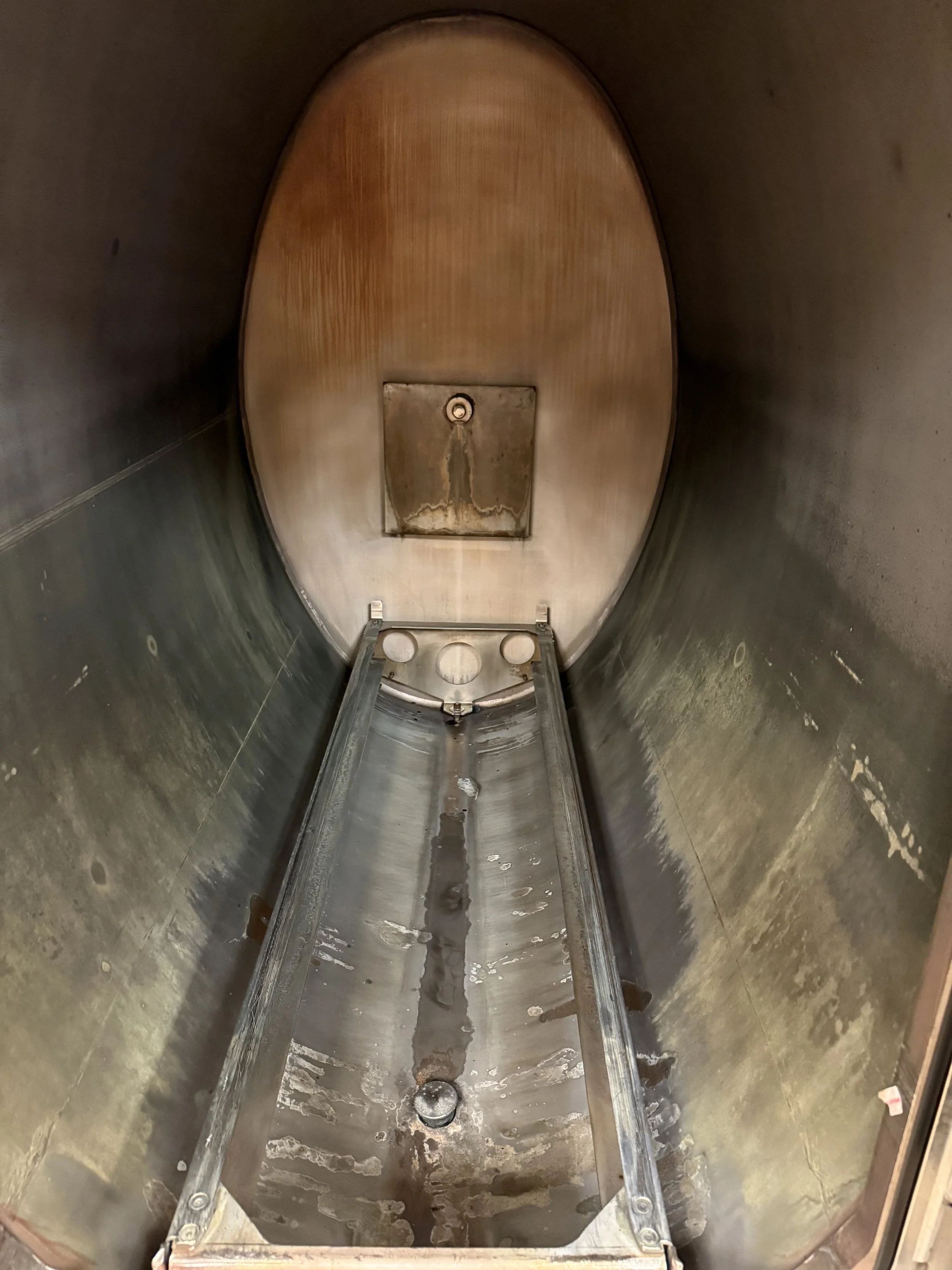Inside a large industrial metal tank or vessel with a rounded interior. The bottom is wet, with some rust, bioburden, and stains. There is a rectangular access panel or hatch at the top end of the tank and metal supports or brackets at the bottom.