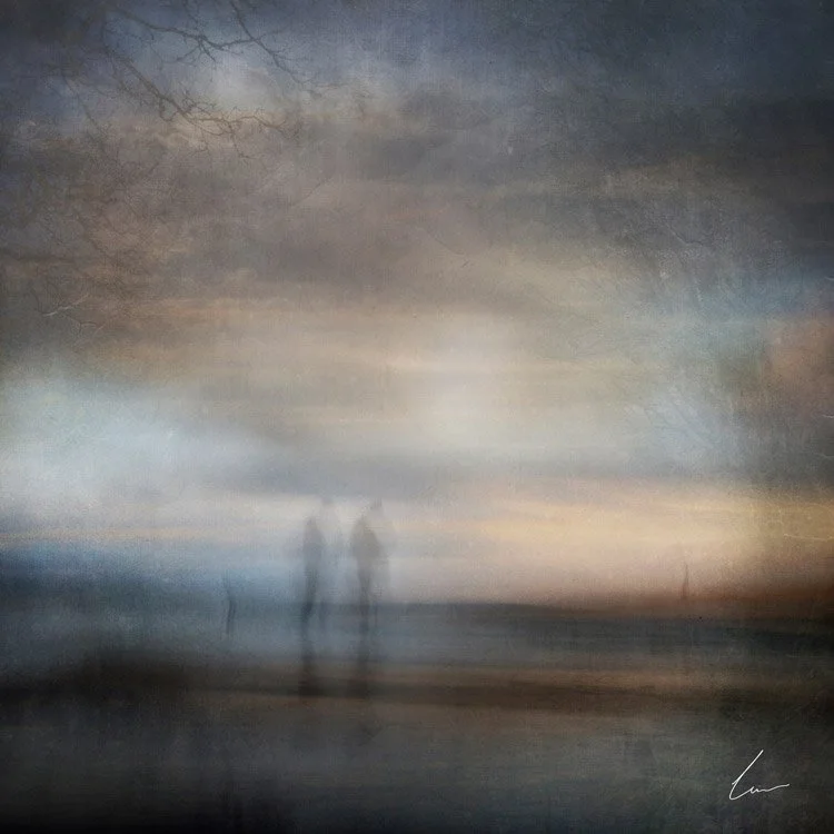 Blurred photograph of two people standing together on a beach at sunset or sunrise with a cloudy sky and bare tree branches in the background.