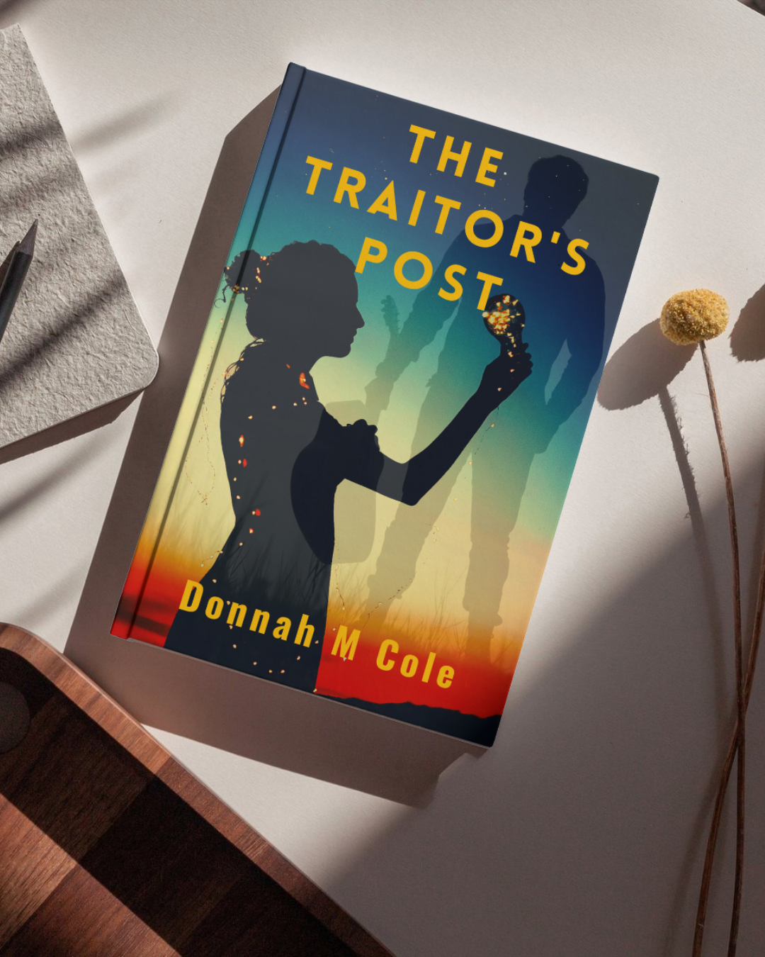 Book titled 'The Traitor's Post' by Donnah M Cole, with silhouette of two people against a colorful sunset background, placed on a white surface with a dried flower and a notepad nearby.