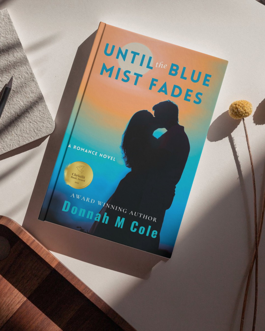 A book titled 'Until the Blue Mist Fades' by Donnah M Cole resting on a white surface with a shadow of a couple embracing on the cover, surrounded by a yellow flower and a gray notebook with a pencil.