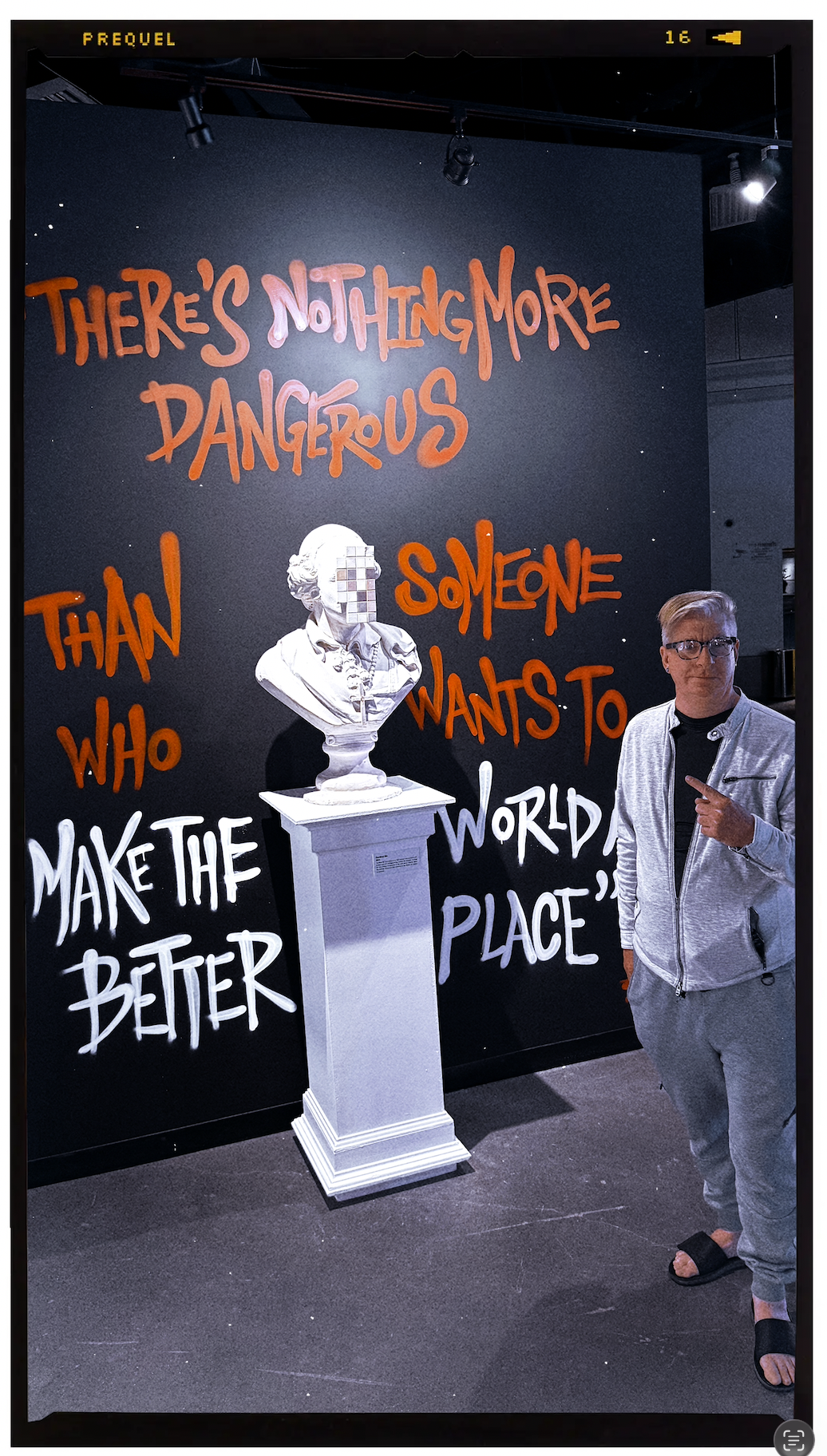 A person standing next to a bust sculpture with a pixelated face, in front of a black wall with red and white handwritten-style text that reads "There’s nothing more dangerous than someone who wants to make the world a better place."