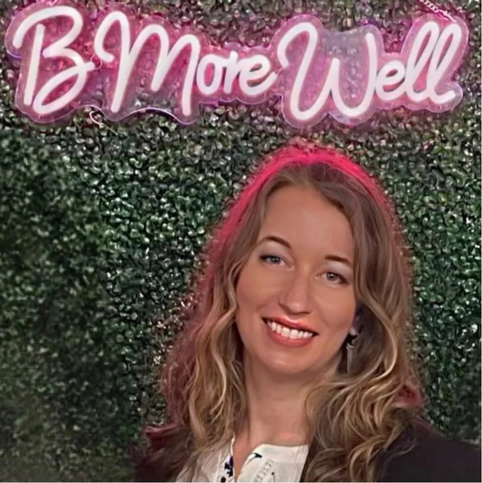A young woman with long wavy blonde hair standing in front of a green hedge wall with a pink neon light sign that reads 'Be More Well'.