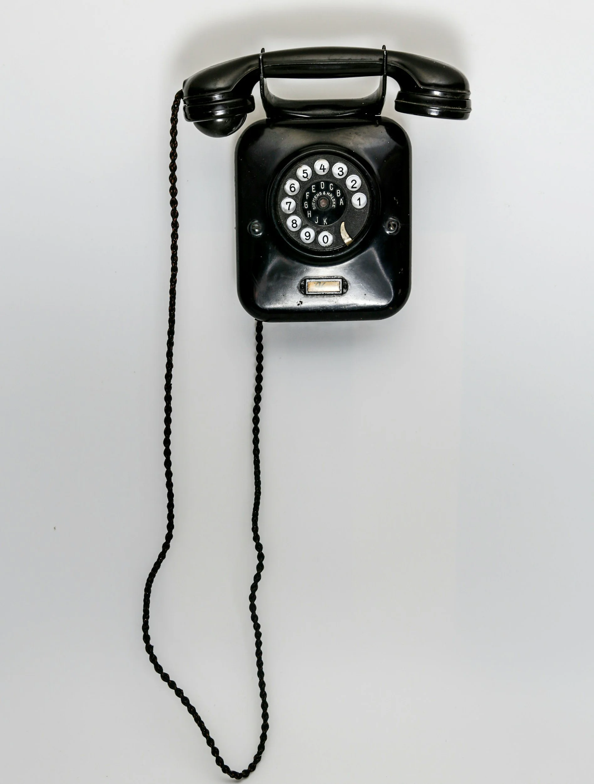 A black rotary dial telephone with a cord attached, mounted on a white wall.