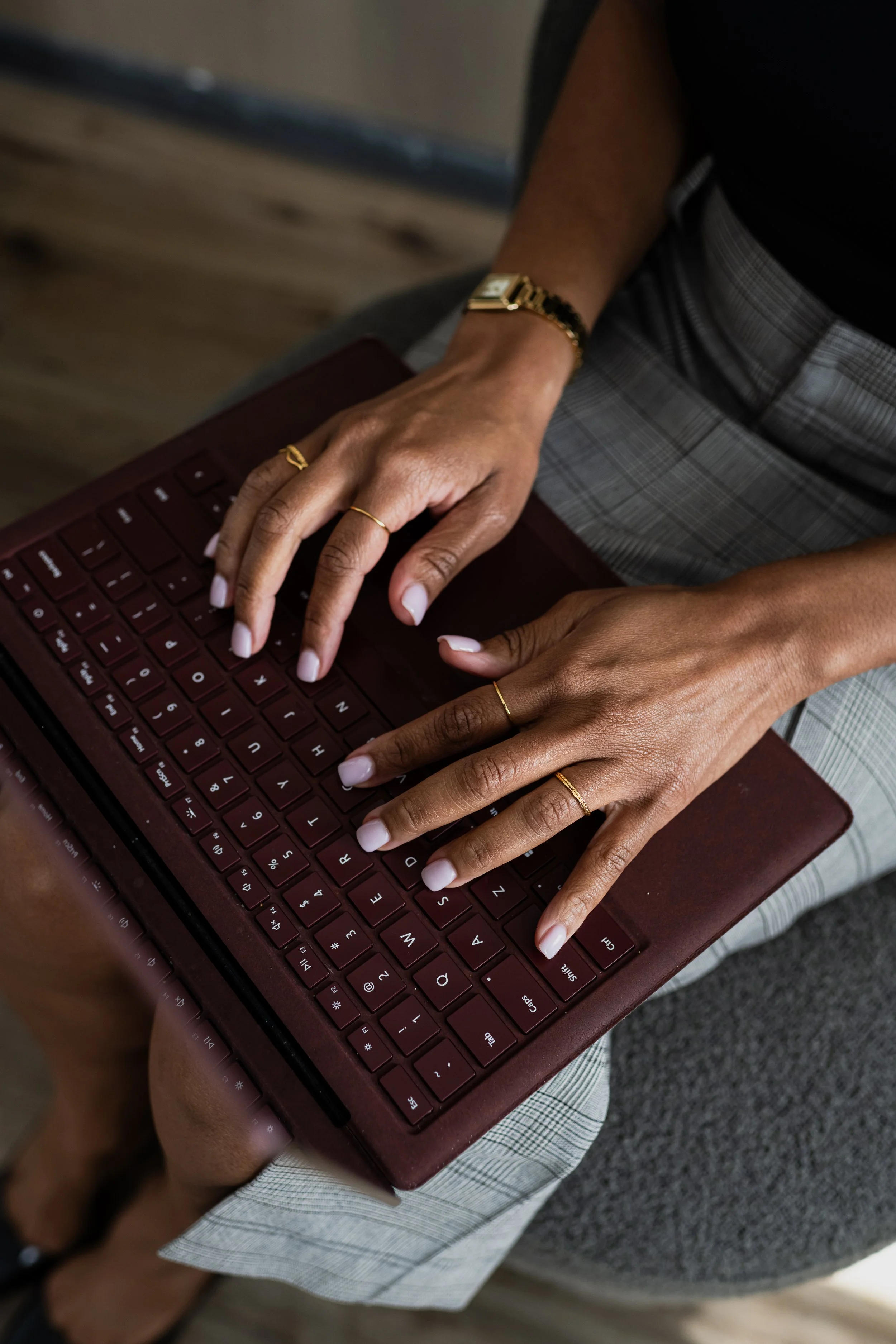 Person sitting cross-legged using a maroon laptop, wearing a gold watch on their wrist and multiple rings on their fingers, dressed in a black top and plaid pants.