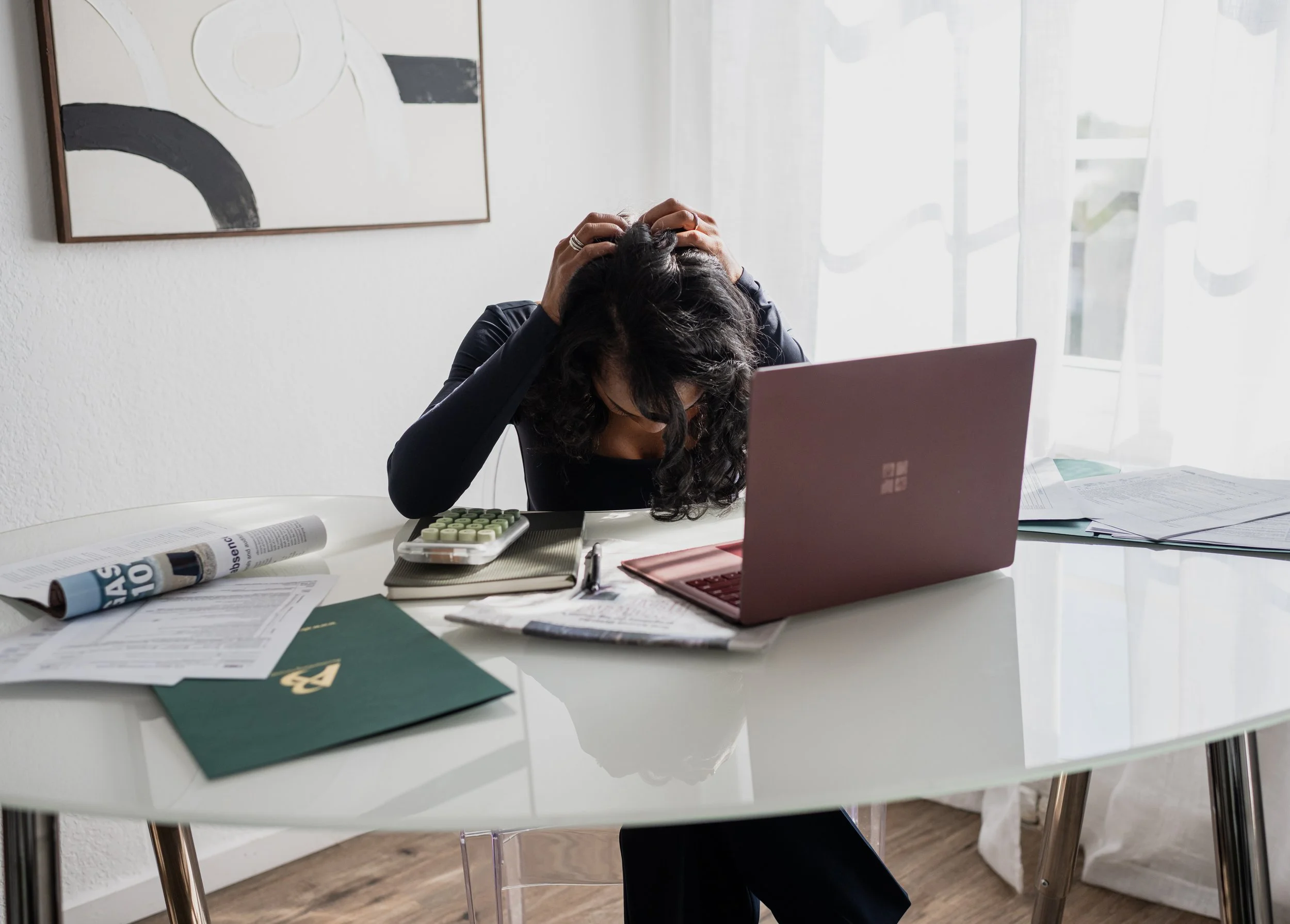 A woman sitting at a cluttered desk, holding her head in frustration, with a laptop, papers, a calculator, and a newspaper on the desk. The background has a white wall and a window with curtains.