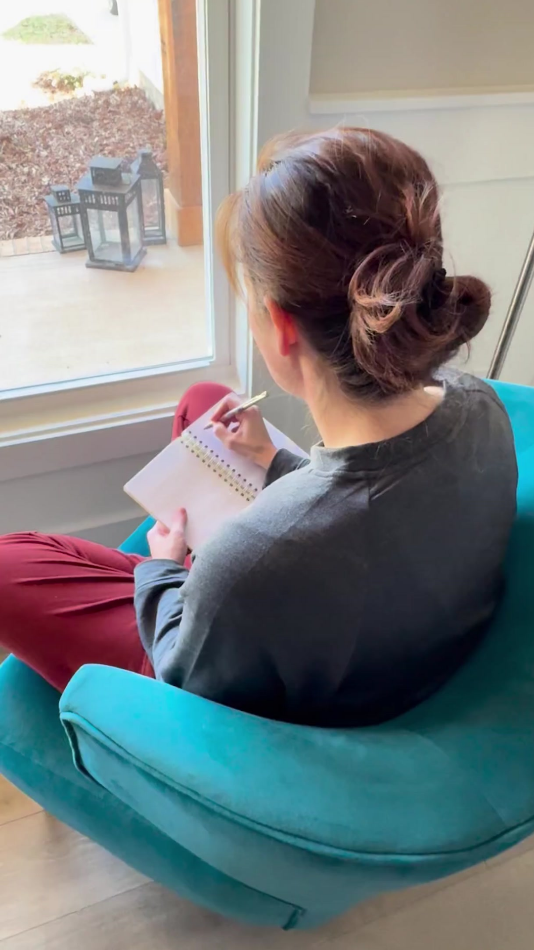 A woman with styled hair sitting on a teal chair, writing in a spiral notebook while looking out a large window.