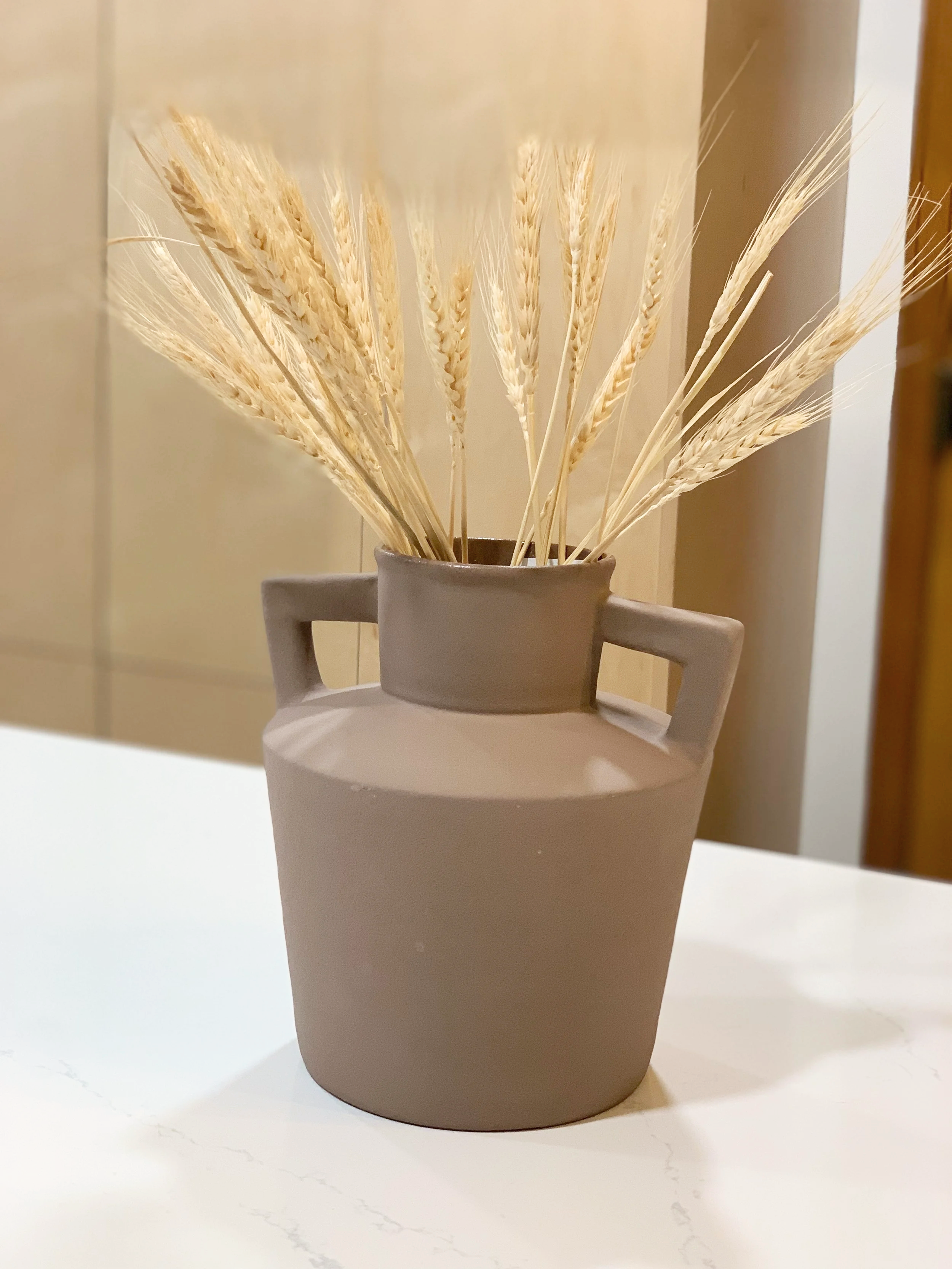 A beige ceramic vase with handles, filled with dried wheat stalks, sits on a white surface.