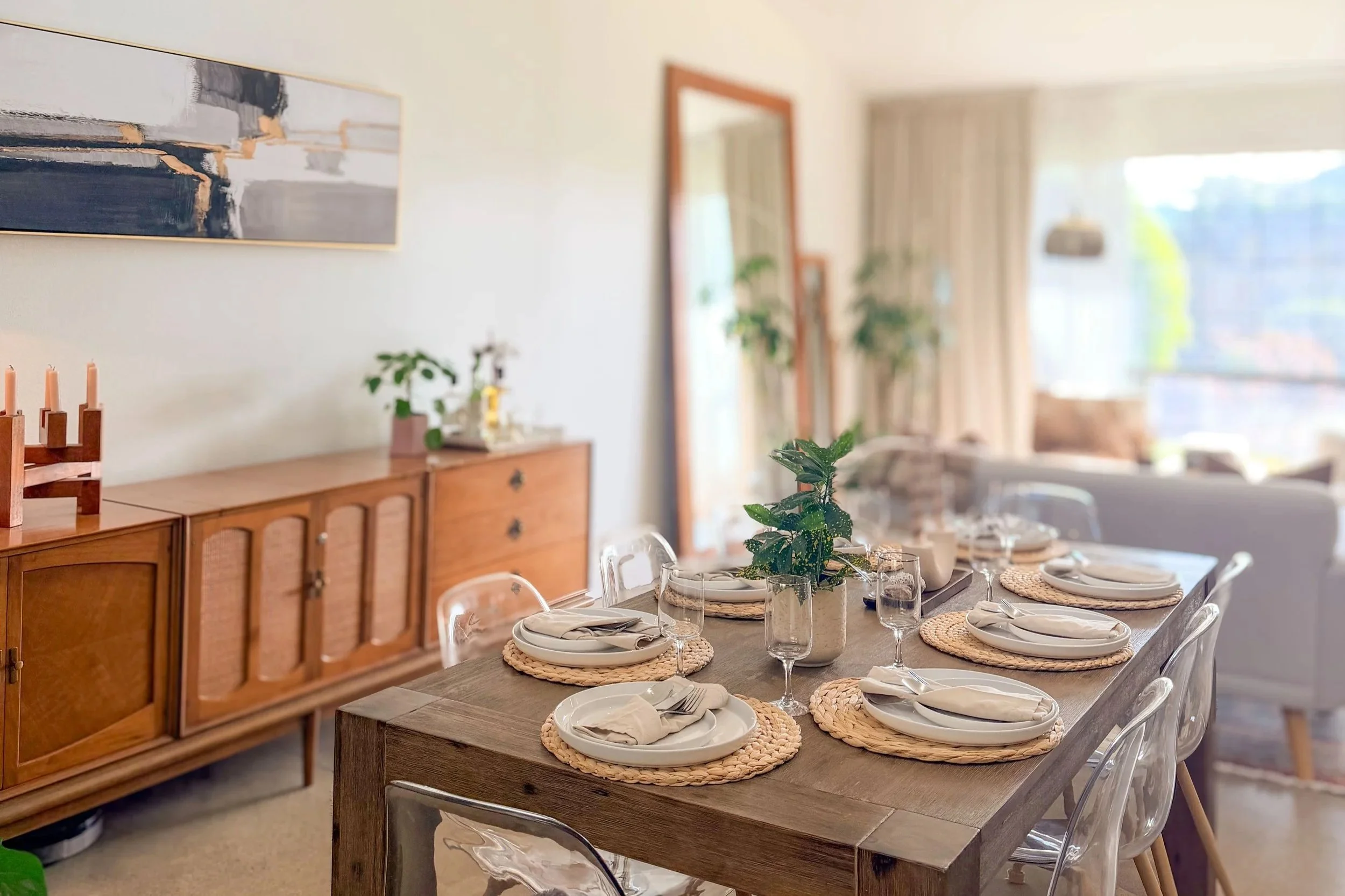 A dining room with a wooden table set with white plates, napkins, glasses, and woven placemats, decorated with a potted plant in the center, and a sideboard with plants, candles, and decorative items against a white wall with artwork. Large window with curtains in the background.