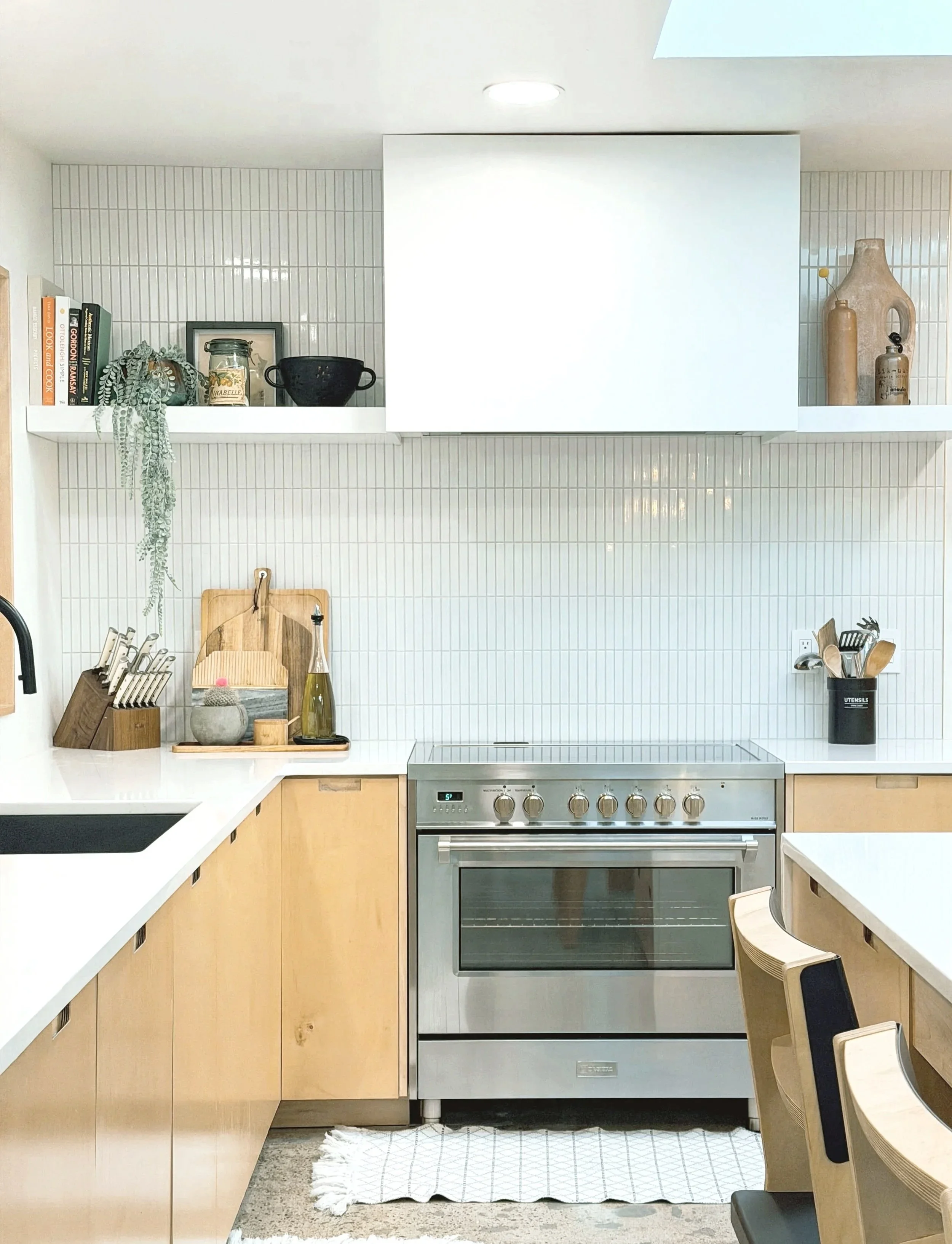 Modern kitchen with light wood cabinets, a stainless steel oven, white countertops, decorative vases, books, plants, and kitchen utensils on a white shelf, with a small woven rug on the floor.