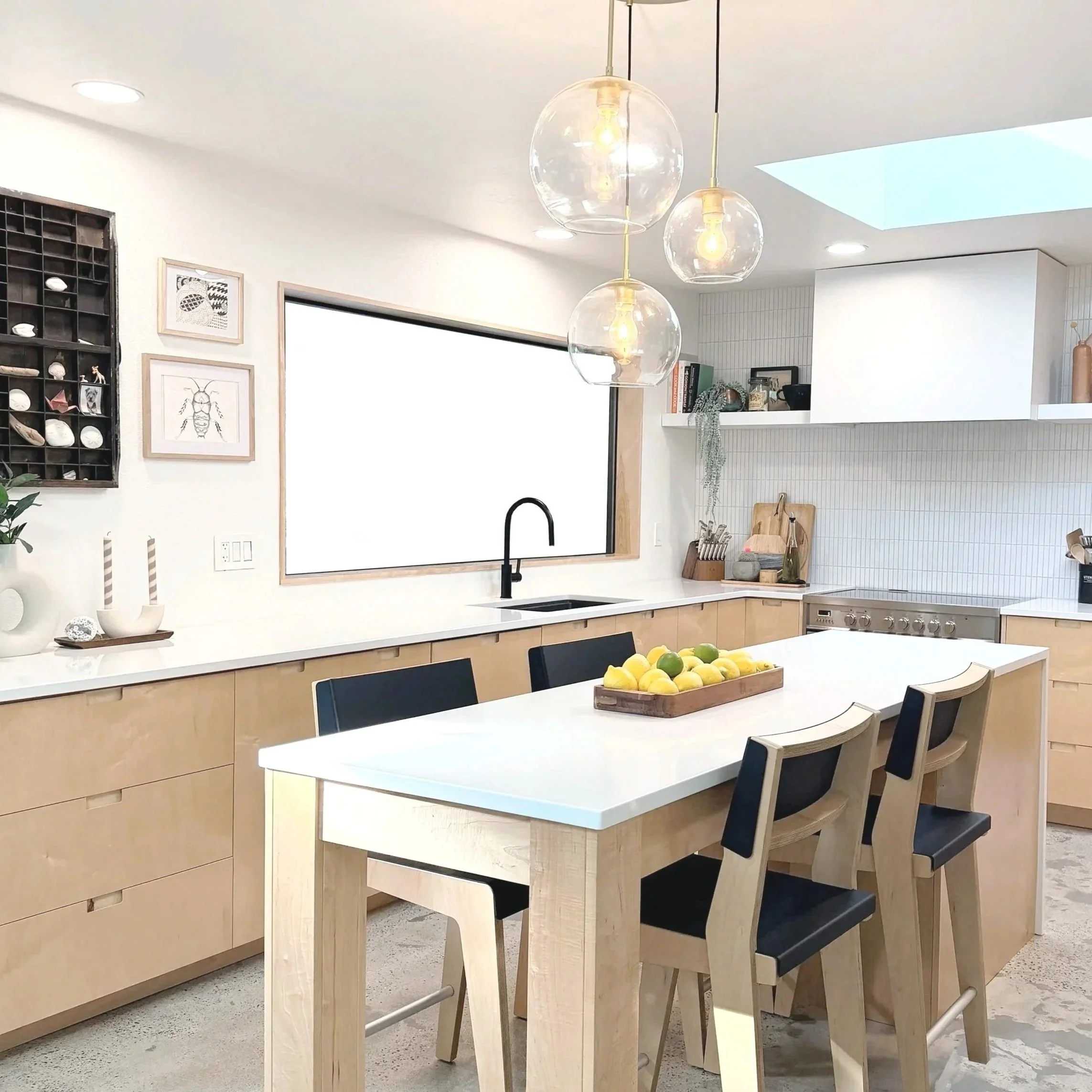 Modern kitchen with white countertops, light wood cabinets, black sink, black chairs around a white island, hanging glass pendant lights, framed art and decor on the wall, open shelves, and a large window.