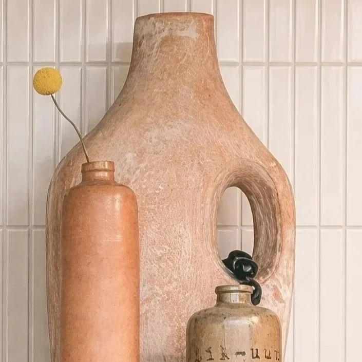 Decorative vases on a tiled wall, including a large ceramic vase with a circular opening, a smaller ceramic vase with a single yellow flower, and a small jar with black stones.