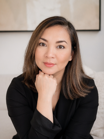 Portrait of a woman with shoulder-length brown hair, wearing a black blazer, resting her chin on her hand, in an indoor setting with a beige wall and artwork in the background.