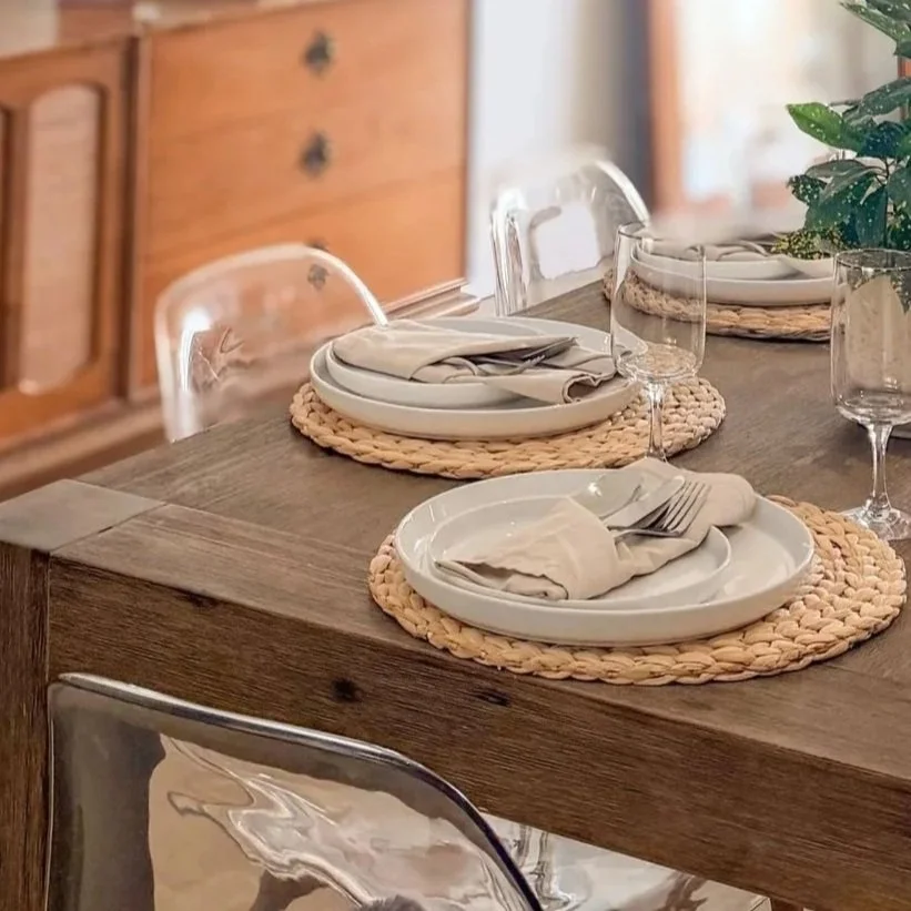 A dining table set with woven placemats, white plates, folded napkins, silverware, and empty wine glasses in a cozy, rustic dining room.
