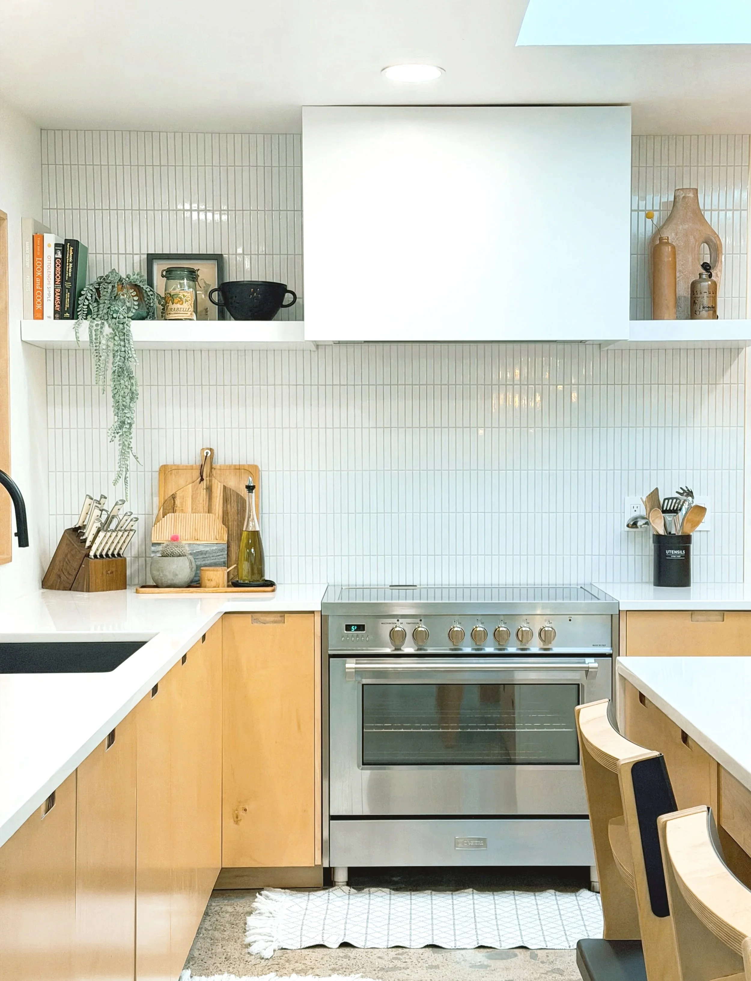 Modern kitchen with light wood cabinets, white countertops, stainless steel oven, and decorative items like books, plants, and pottery on shelves.