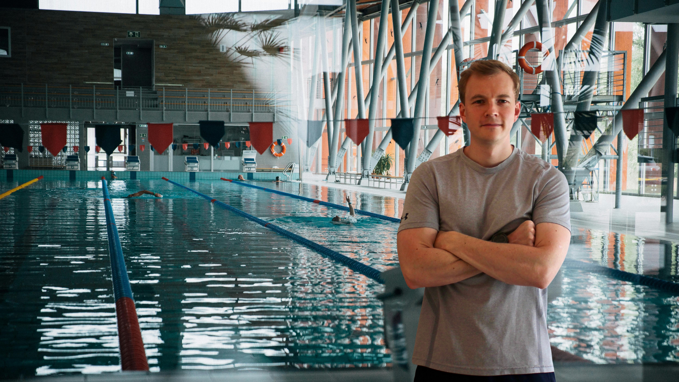 A man with crossed arms standing in front of an indoor swimming pool with swimmers practicing laps.