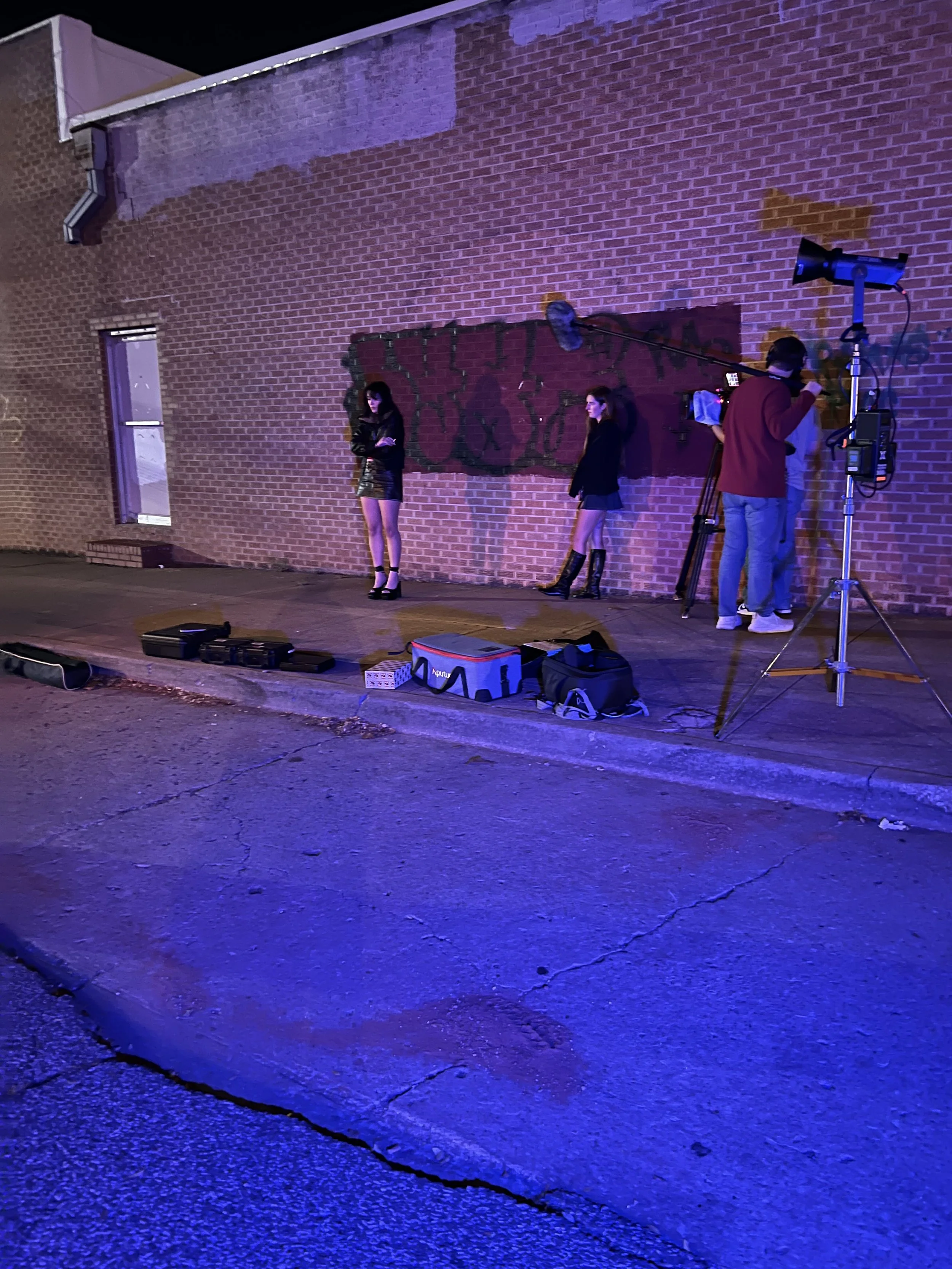 The 'Call Girl' (2026) film crew standing on the street, filming actresses' Conner Cagle and Molly Funderburk on a city sidewalk at night, with film equipment and a brick building backdrop.