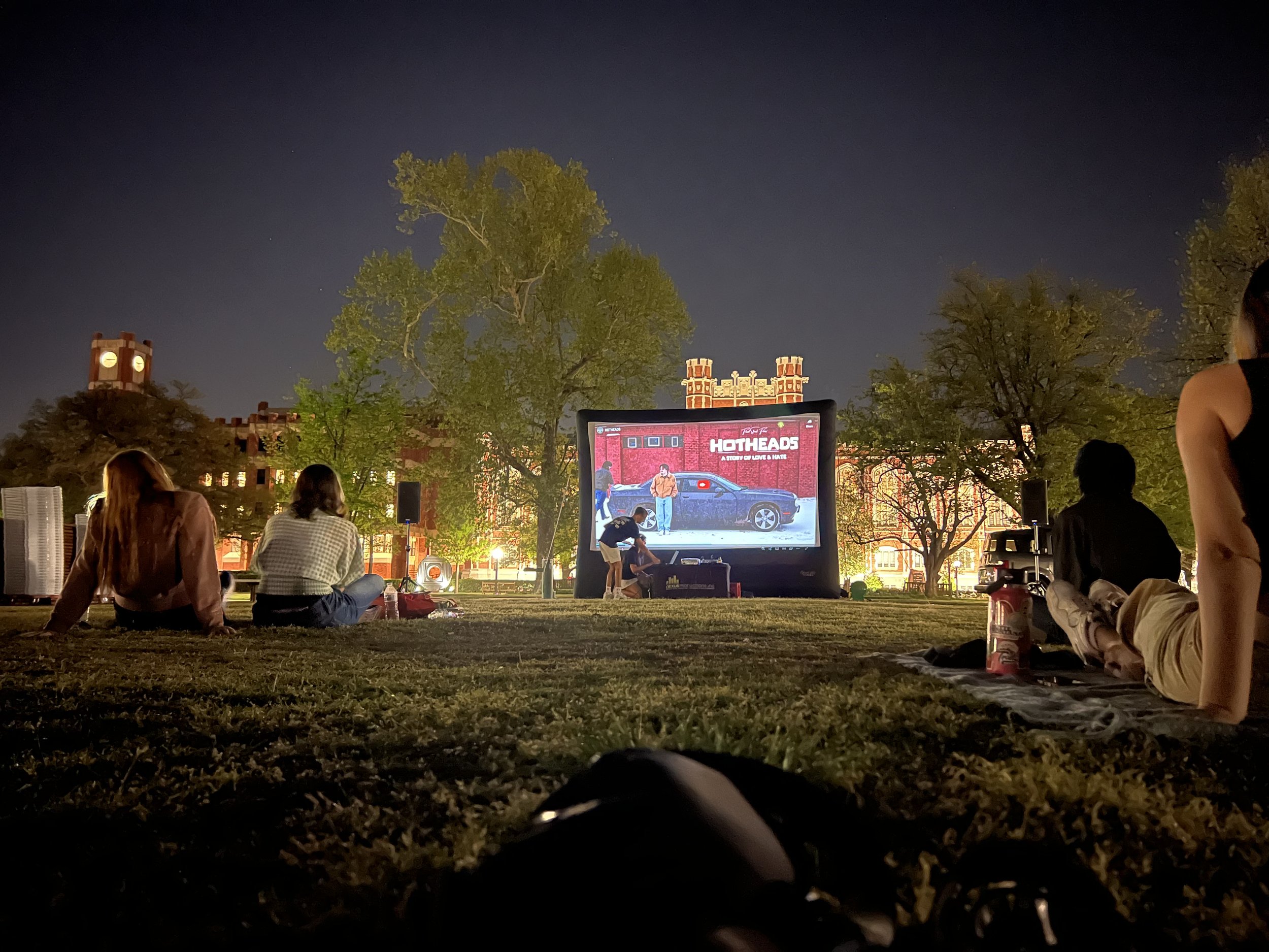 People sitting on the grass watching a movie outdoors at night, with a large screen displaying the movie and the words 'HOTHEADS', Directed by Ryan Christopher Park & Jake Ayers.
