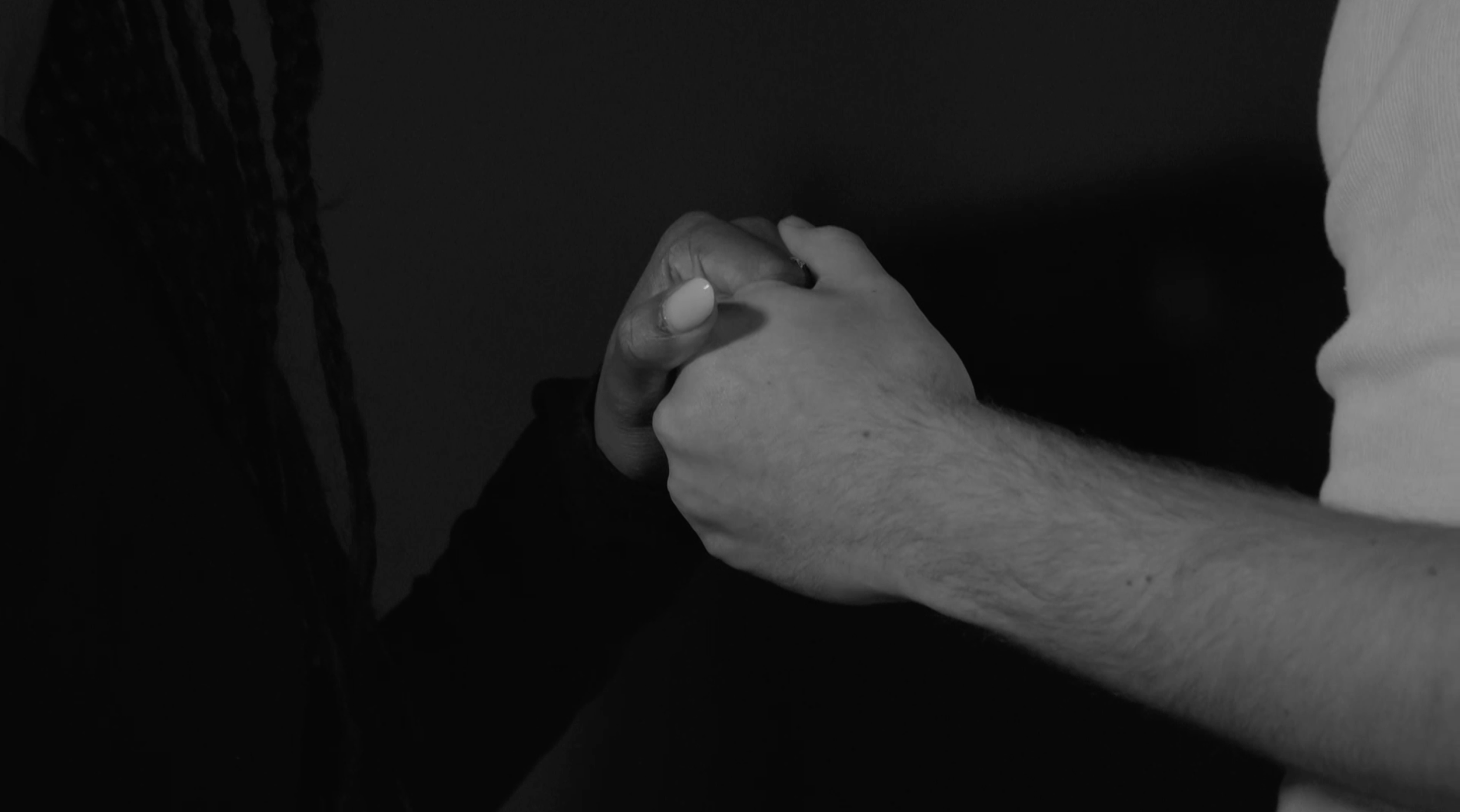 Close-up of two people holding hands, one with a darker complexion and the other with a lighter complexion, in a tender embrace. Still from the short film, THE IRON BULL (2024).