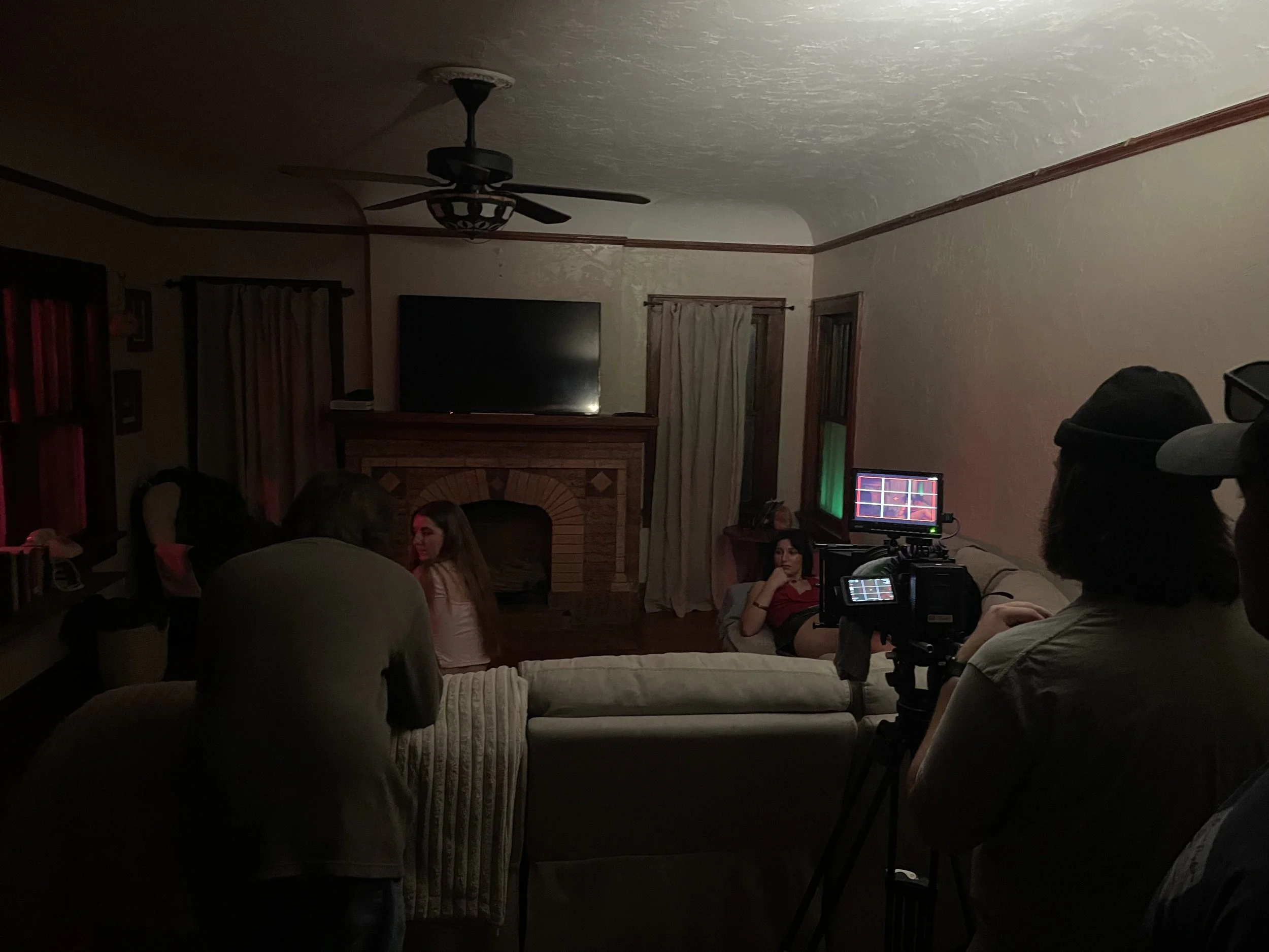 From 'Call Girl' (2025), the crew is filming a scene in a dimly lit living room with a fireplace, a television above it, and a woman sitting on a couch in a red shirt.