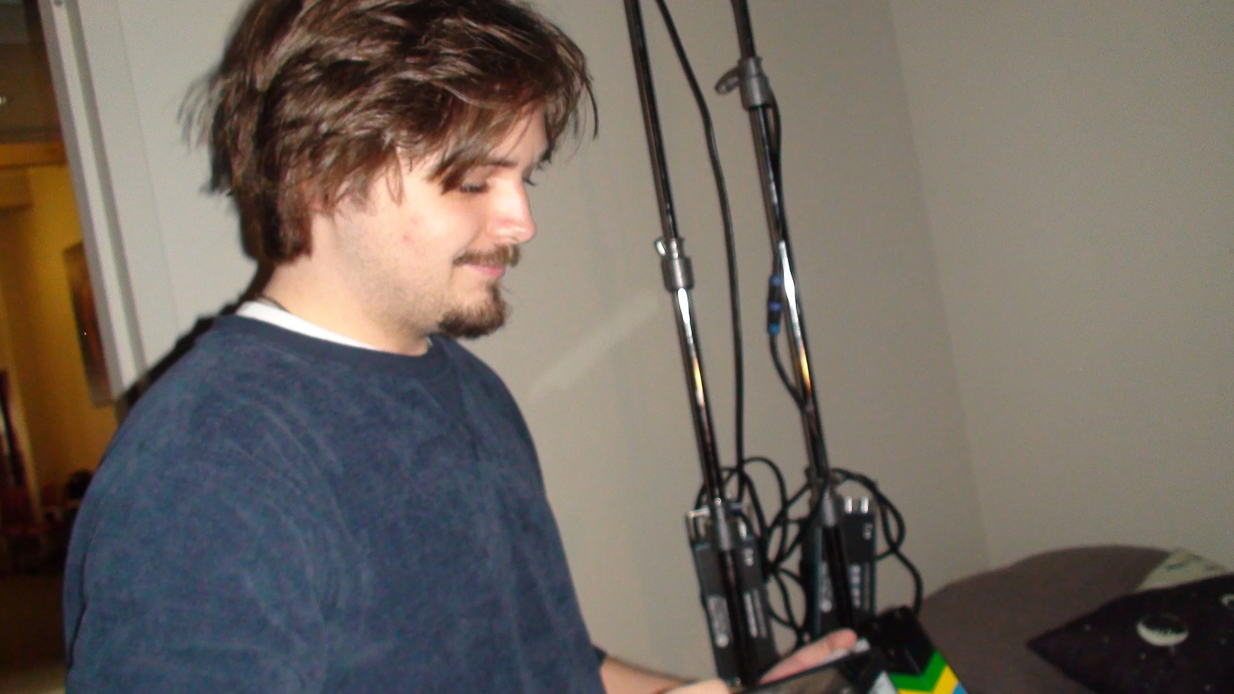Ryan Christopher Park looking down, standing on the set of 'Name' (2025) next to a bed with a pillow and a lightstand, in a room with a beige wall and some black electronic equipment with cables hanging on the wall