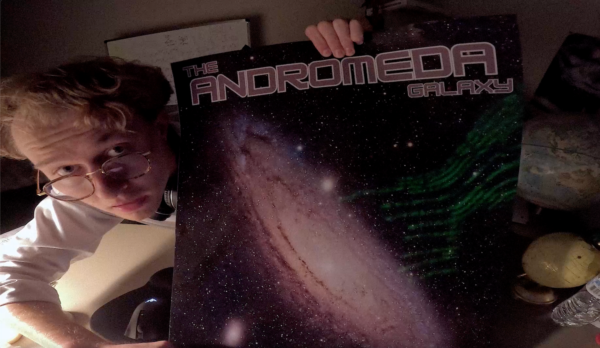 Jacob Leo Marino in a still from NAME (2026), holding up a poster of a galaxy with the title 'The Andromeda Galaxy' in purple lettering.