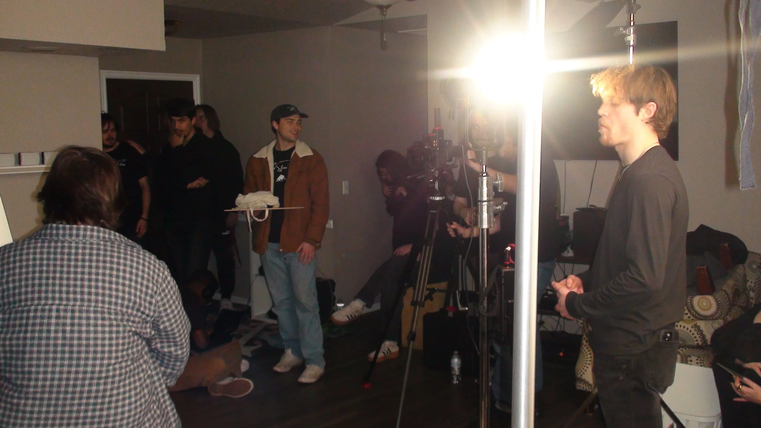 A film or video production scene with a group of people, including a director and camera crew, preparing for a shoot. Bright lighting equipment is visible, and the target person is standing in front of the camera.