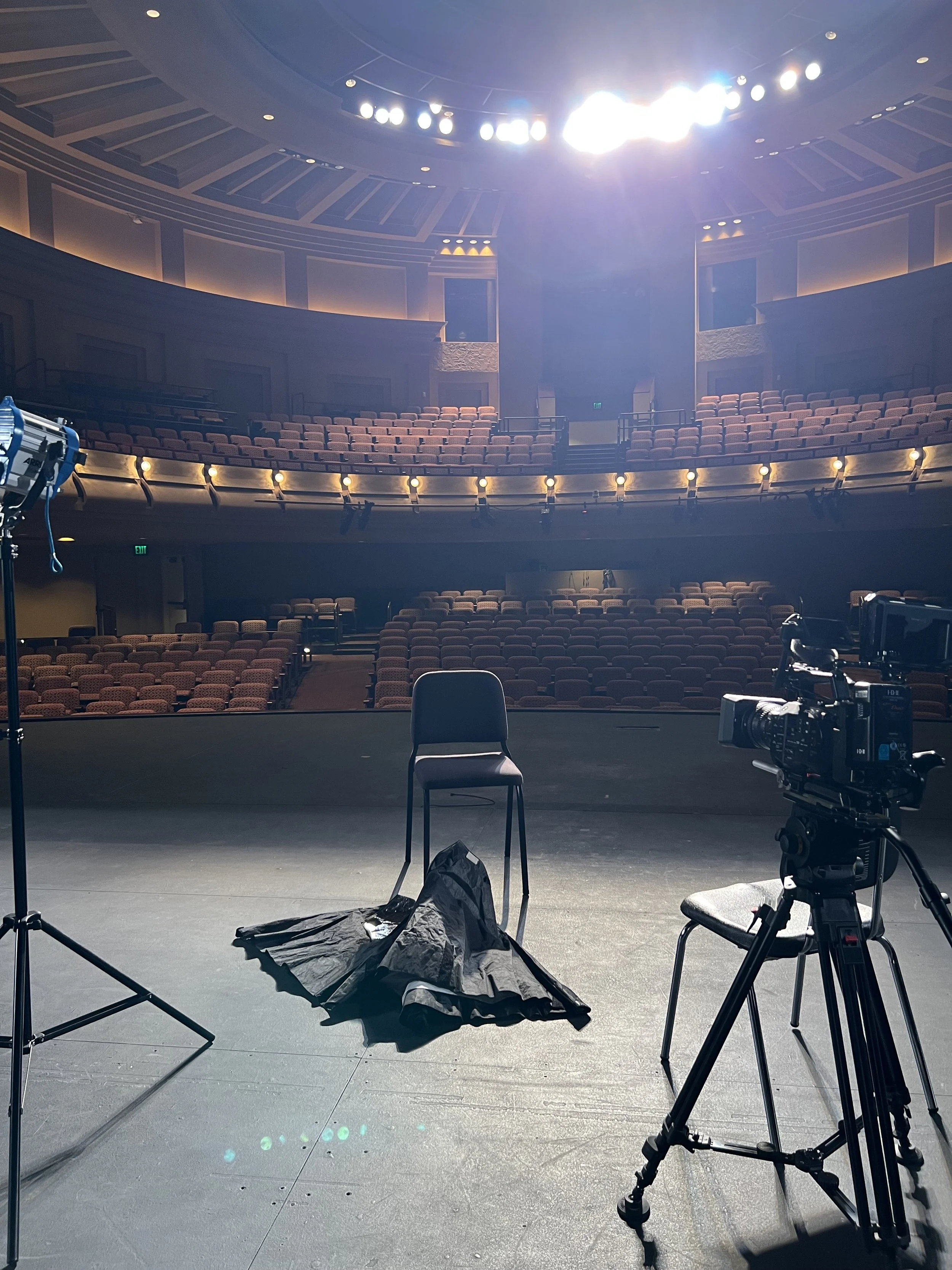 An empty theater stage with a chair, a black cloth, a camera on a tripod, and lighting equipment, facing an audience seating area with rows of empty seats in a large auditorium.