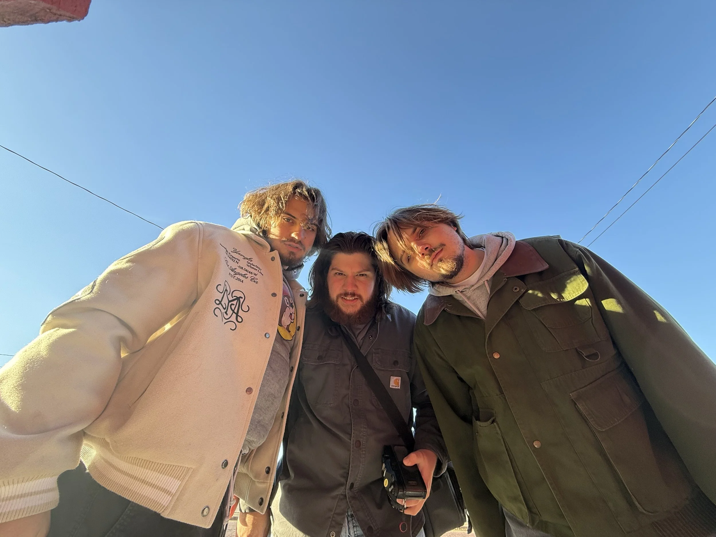 Nathan Lowenstein, Ryan Christopher Park, and Jake Ayers looking down at the camera, outside on a clear day with a blue sky. They are dressed in casual jackets and hoodies.