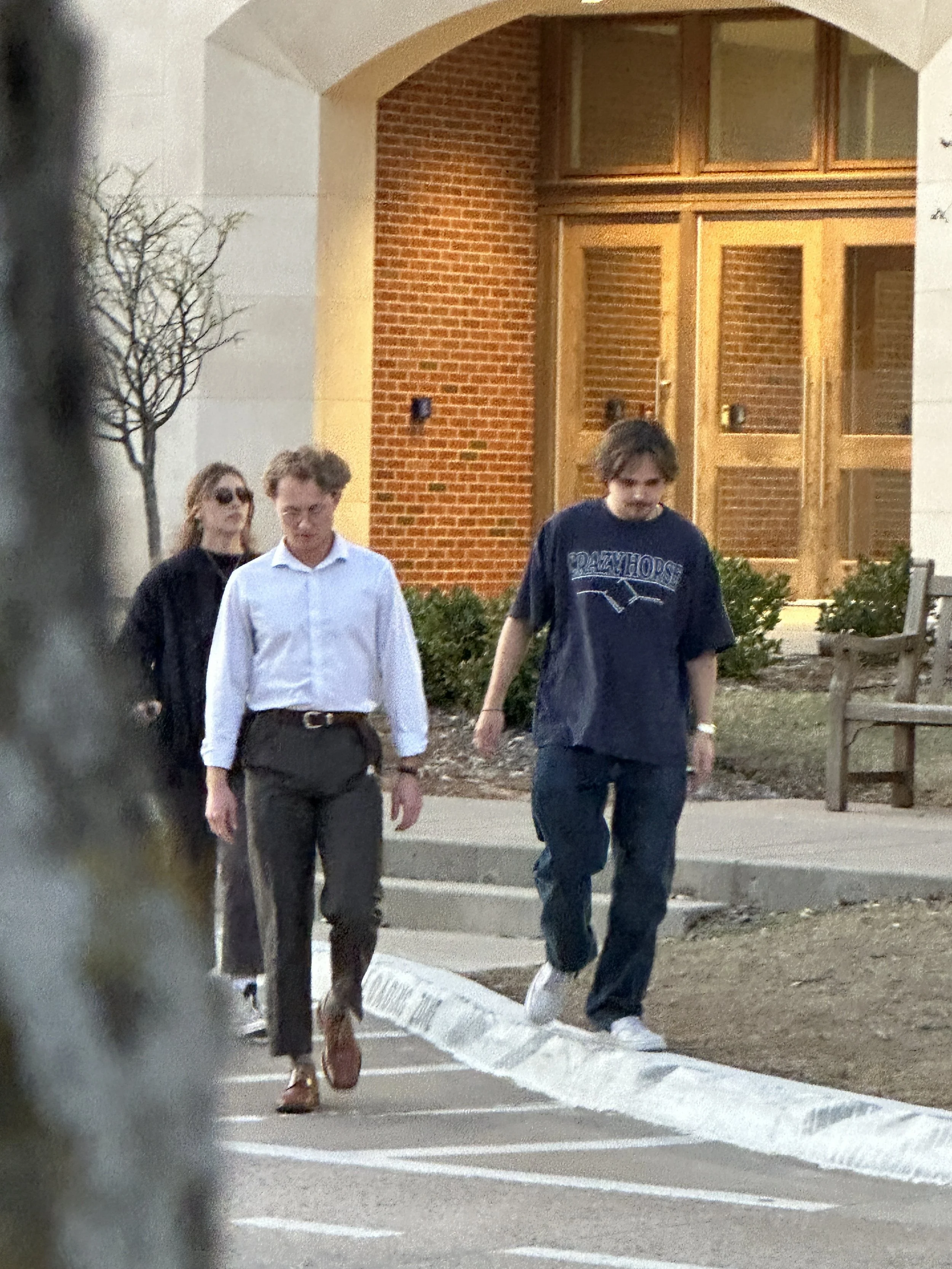 Three people walking on a sidewalk outside a building with a brick and wood door, from the set of 'Name' (2025).