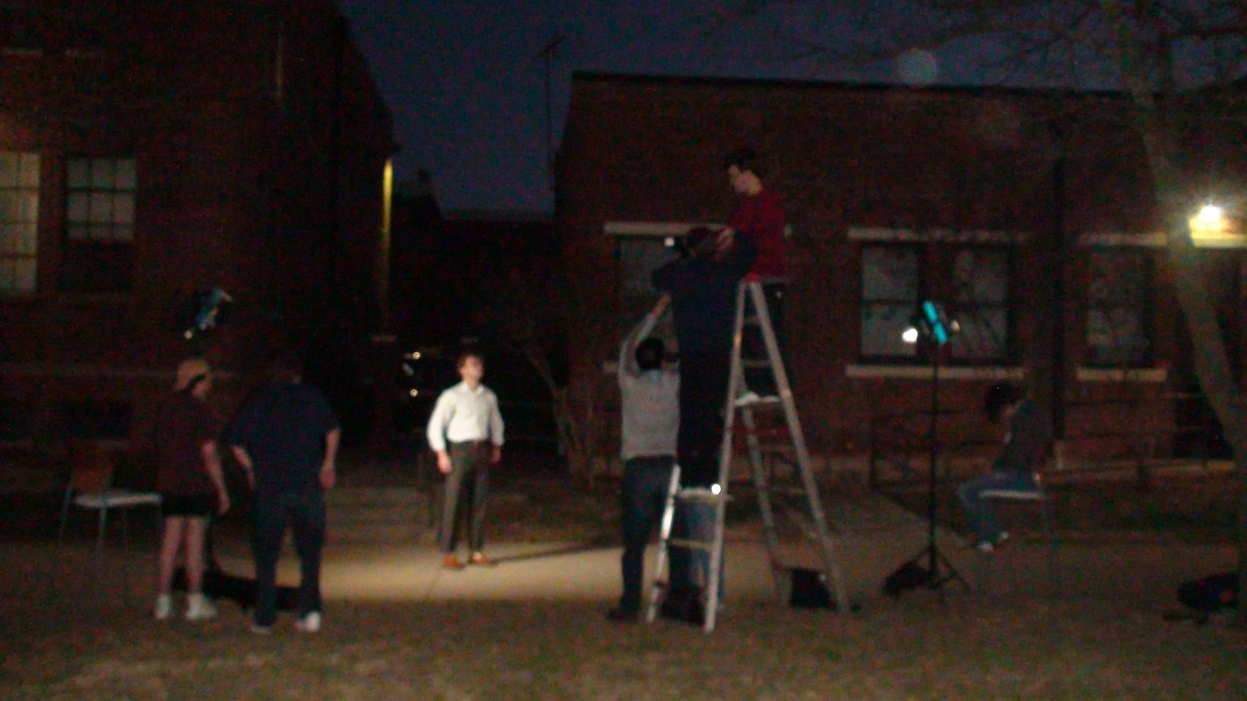 Behind the scenes photo from 'Name' (2025) of the film crew setting up an integral scene outside at night, including the camera team climbing a ladder, lights, and an actor being photographed from the ladder in front of a brick building.