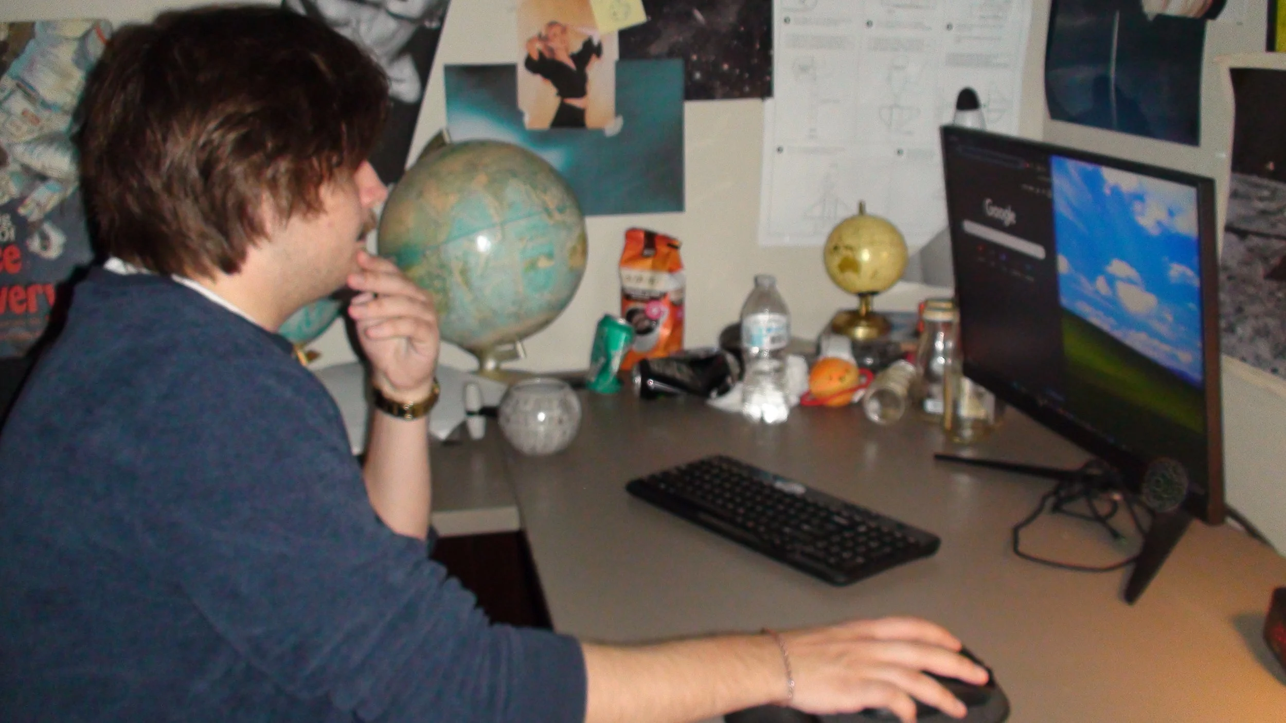 Ryan Christopher Park sitting at cluttered desk in front of a computer monitor, with various objects including globes, bottles, trash, and papers on the desk and wall, on the set of his short film 'Name' (2025).