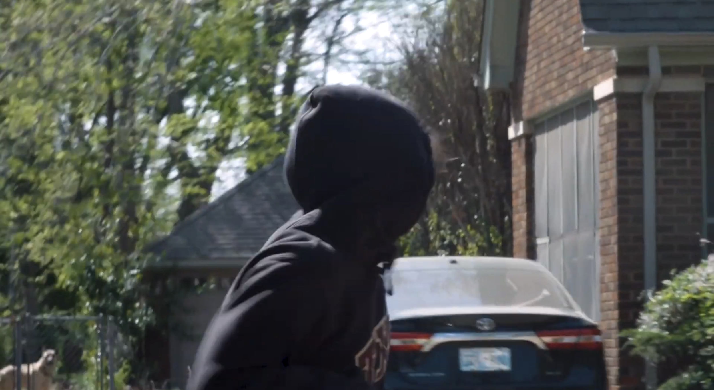 A person wearing a black hoodie is standing outdoors near a car with a license plate, in a residential area with brick houses and trees. Still from the short film, THE IRON BULL (2024).