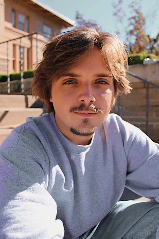 Ryan Christopher Park. A young man with light brown hair, a mustache, and a goatee taking a selfie outdoors on a sunny day, sitting on steps in front of a brick building with trees in the background.