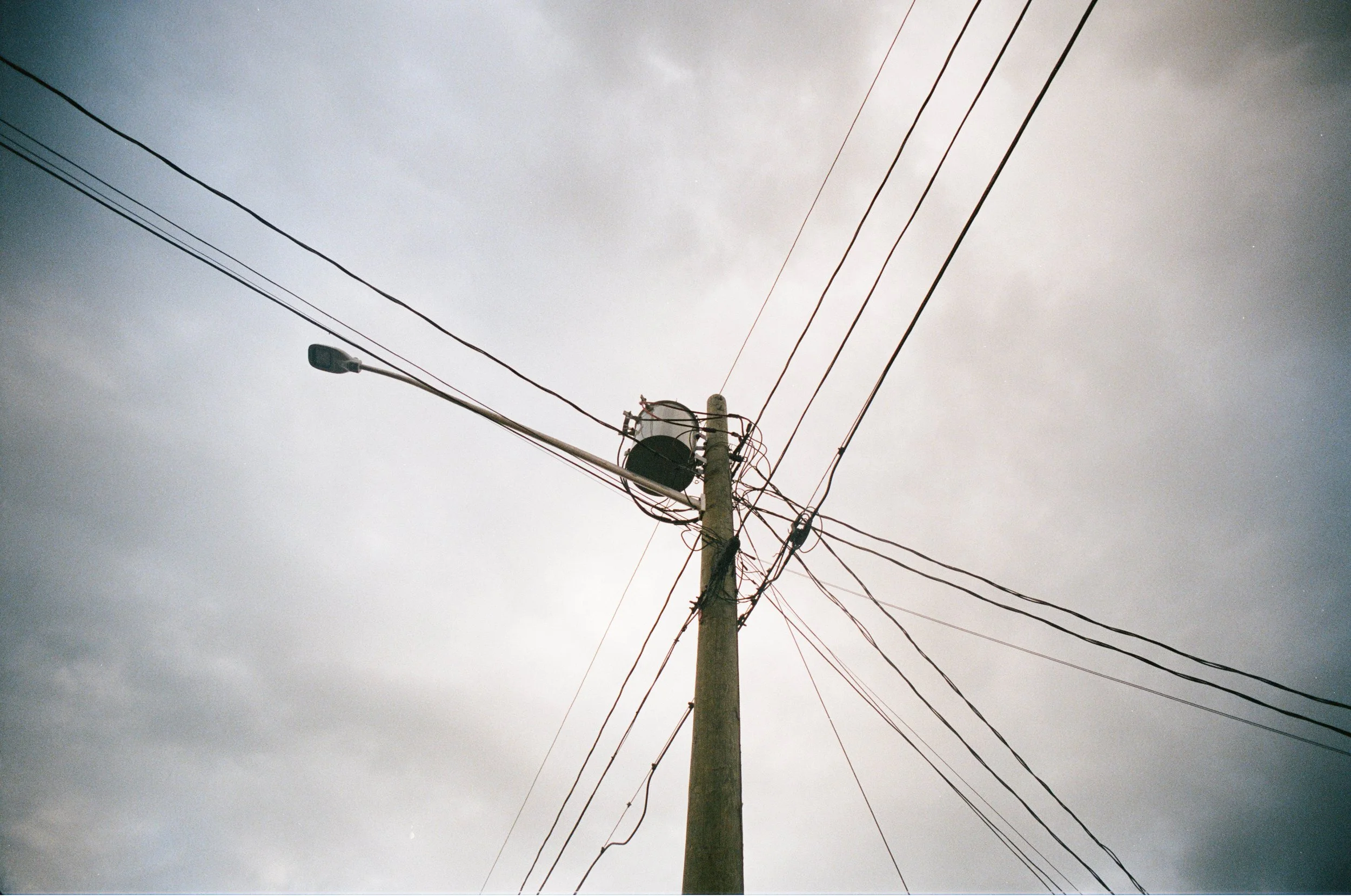 A wooden utility pole with multiple electrical wires extending in various directions, with a streetlight attached, under a cloudy sky. Film photograph by Jake Ayers.