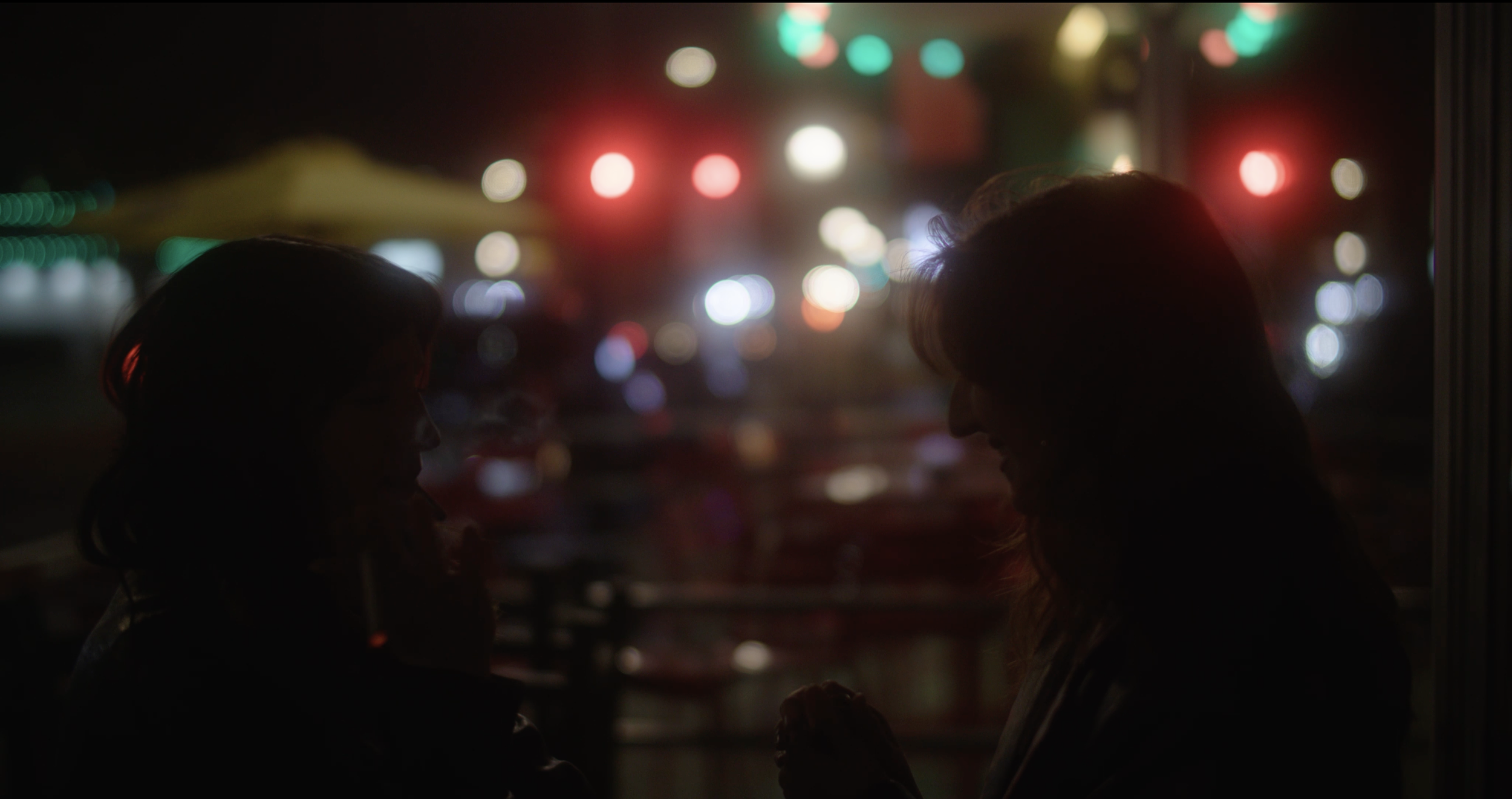 Silhouettes of two women talking inside a dimly lit place with colorful city lights visible through the window at night. Still from the short film CALL GIRL (2026).