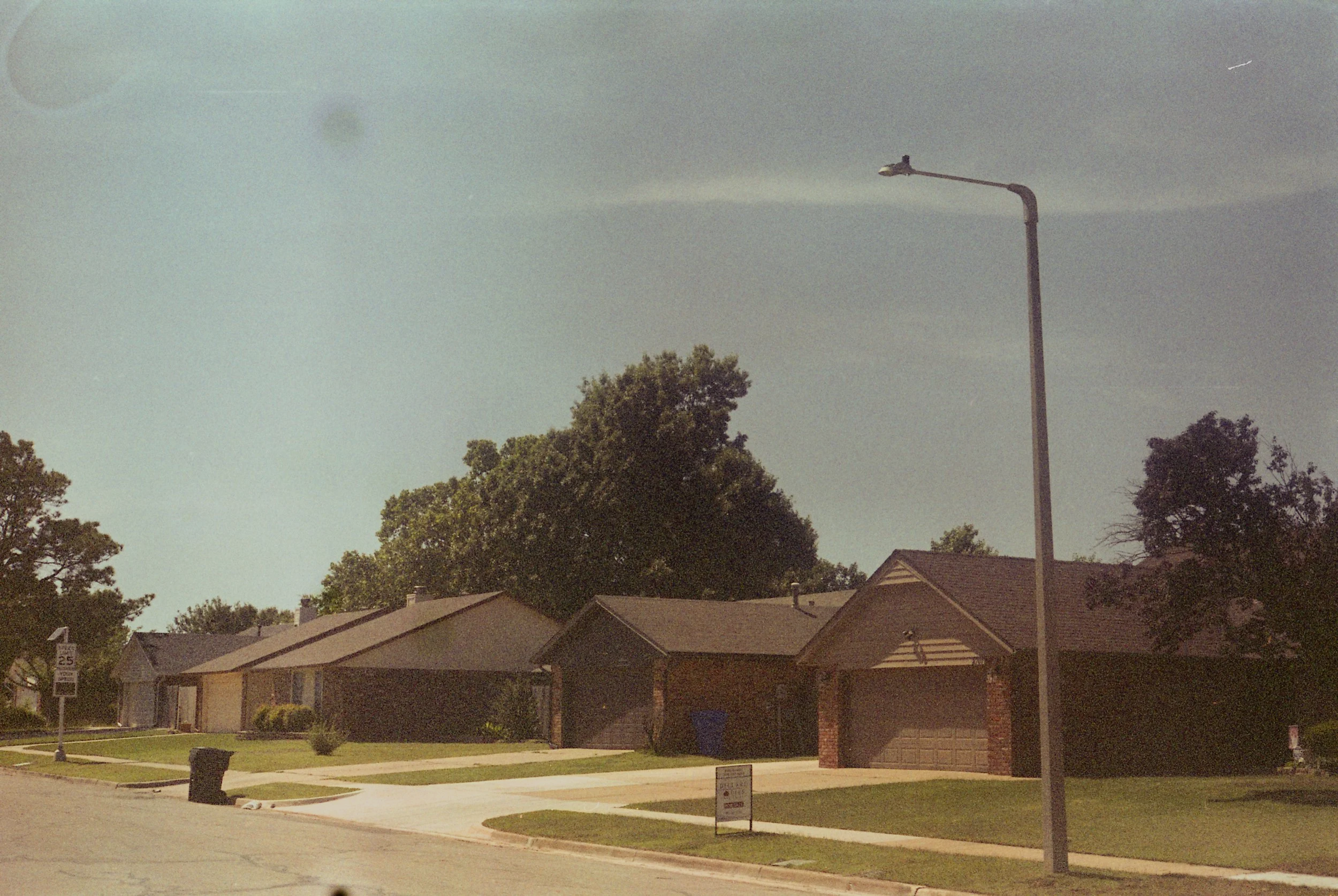A suburban street with single-family homes, a streetlamp, a trash bin, and a sign, with a clear sky and trees in the background. Film photo taken by Jake Ayers.