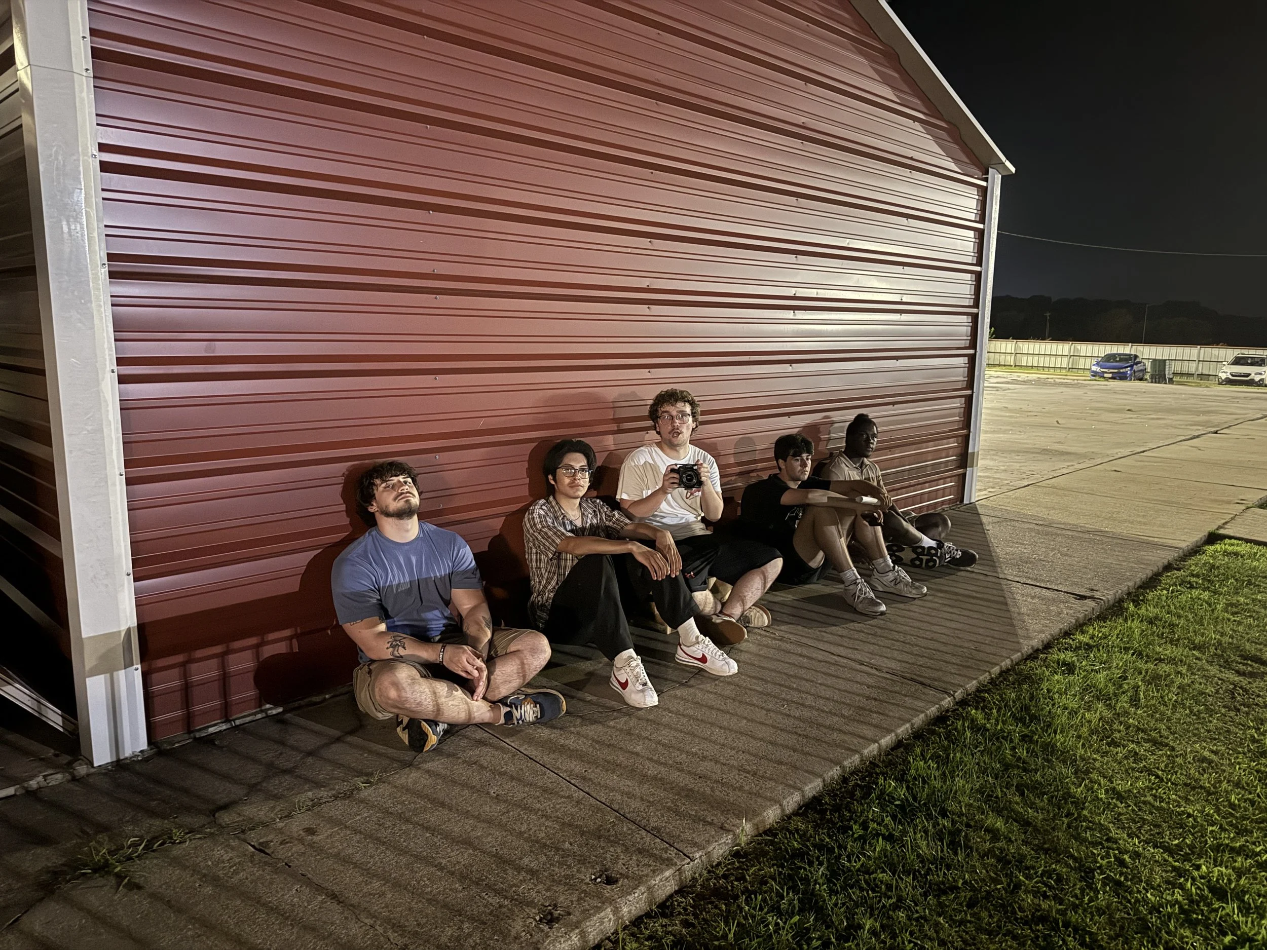 Five young men sitting against a red metal building at night, with a parking lot and cars visible in the background.