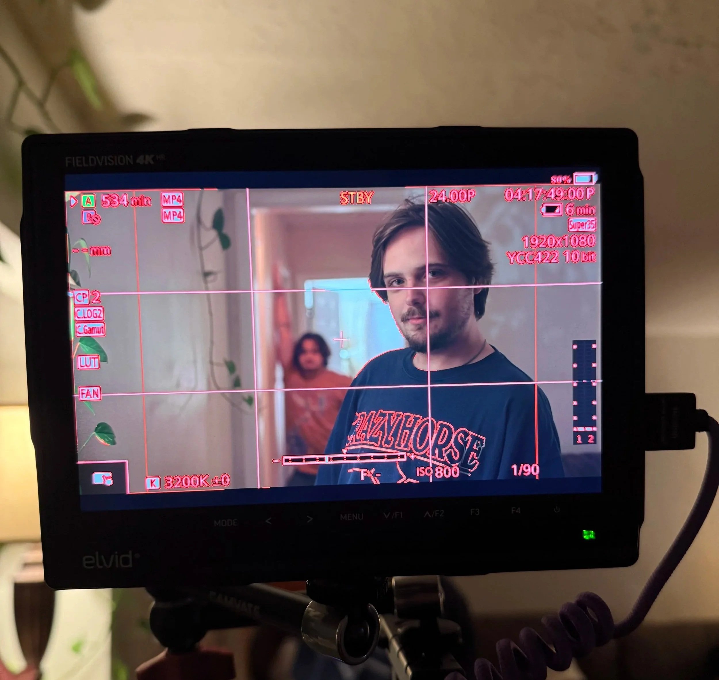On a camera monitor, Ryan Christopher Park is looking at the camera, wearing a dark T-shirt with red and white text. In the background, Nathan Lowenstein with curly hair and a beige shirt is standing in a brightly lit corridor.